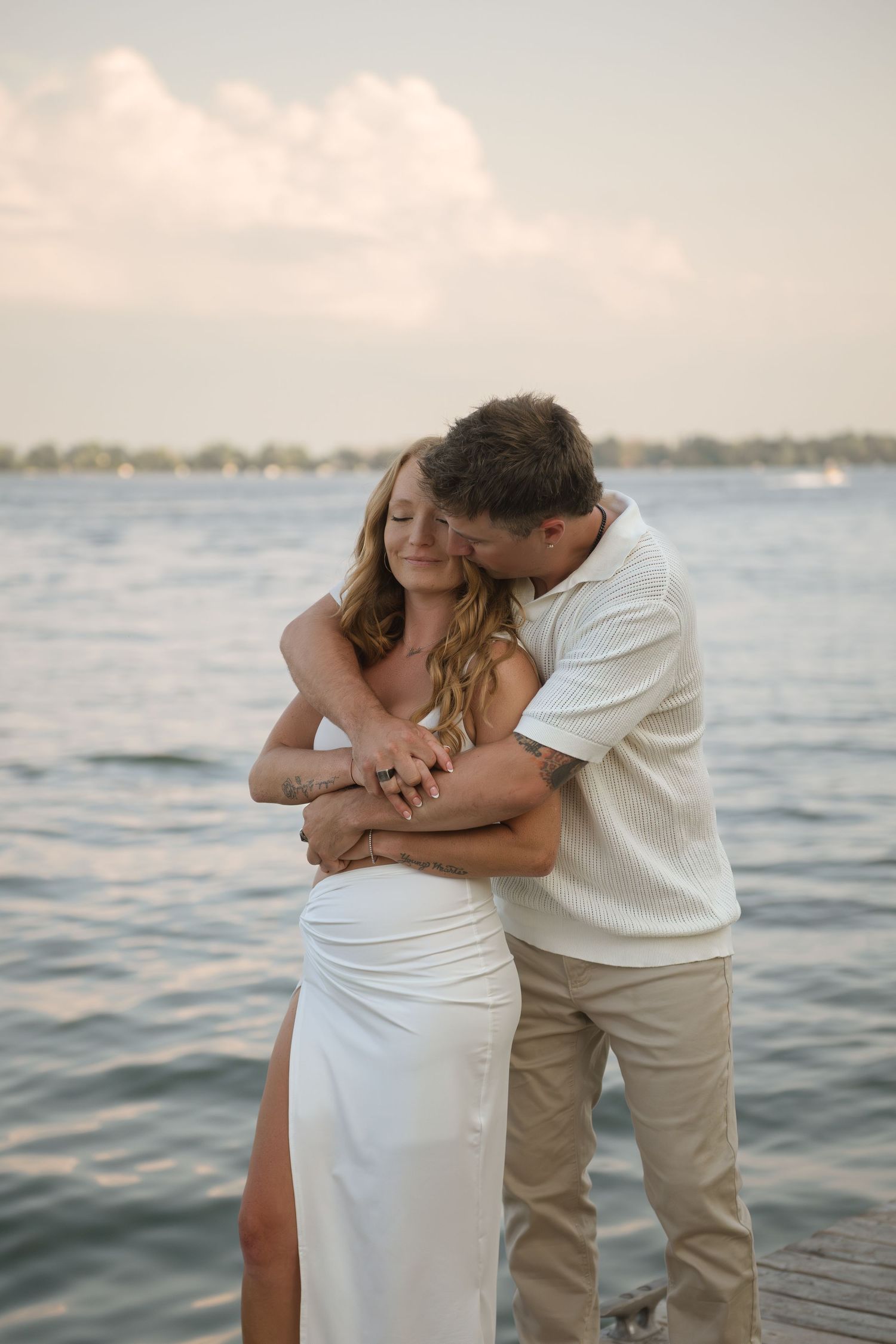 Loving couple embraces on dock with serene water and sky backdrop.