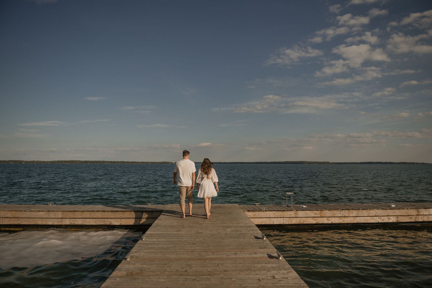 A couple stands together at the end of a wooden dock overlooking a serene lake.