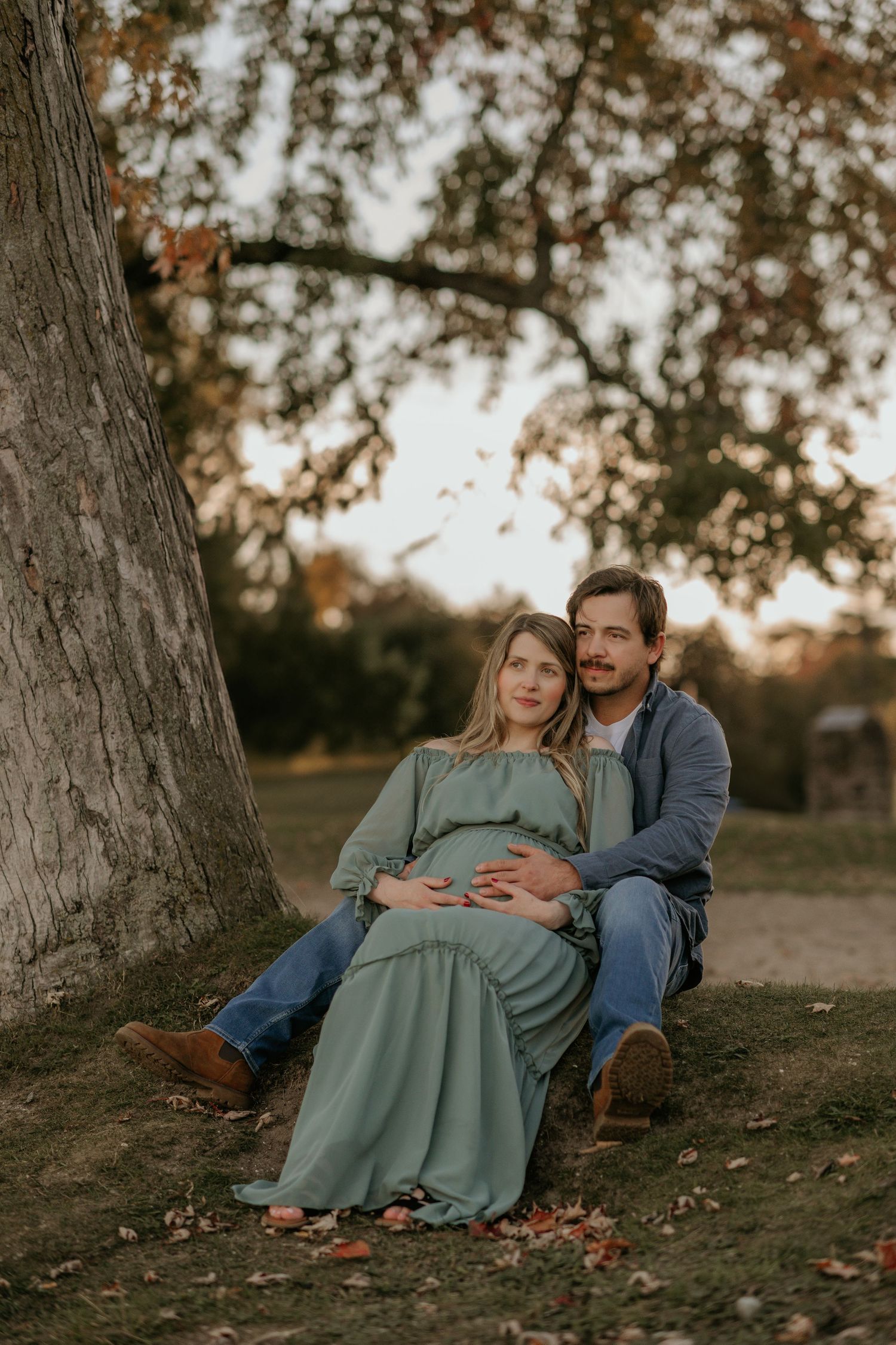 Couple rests against tree during sunset maternity session in mint green dress.