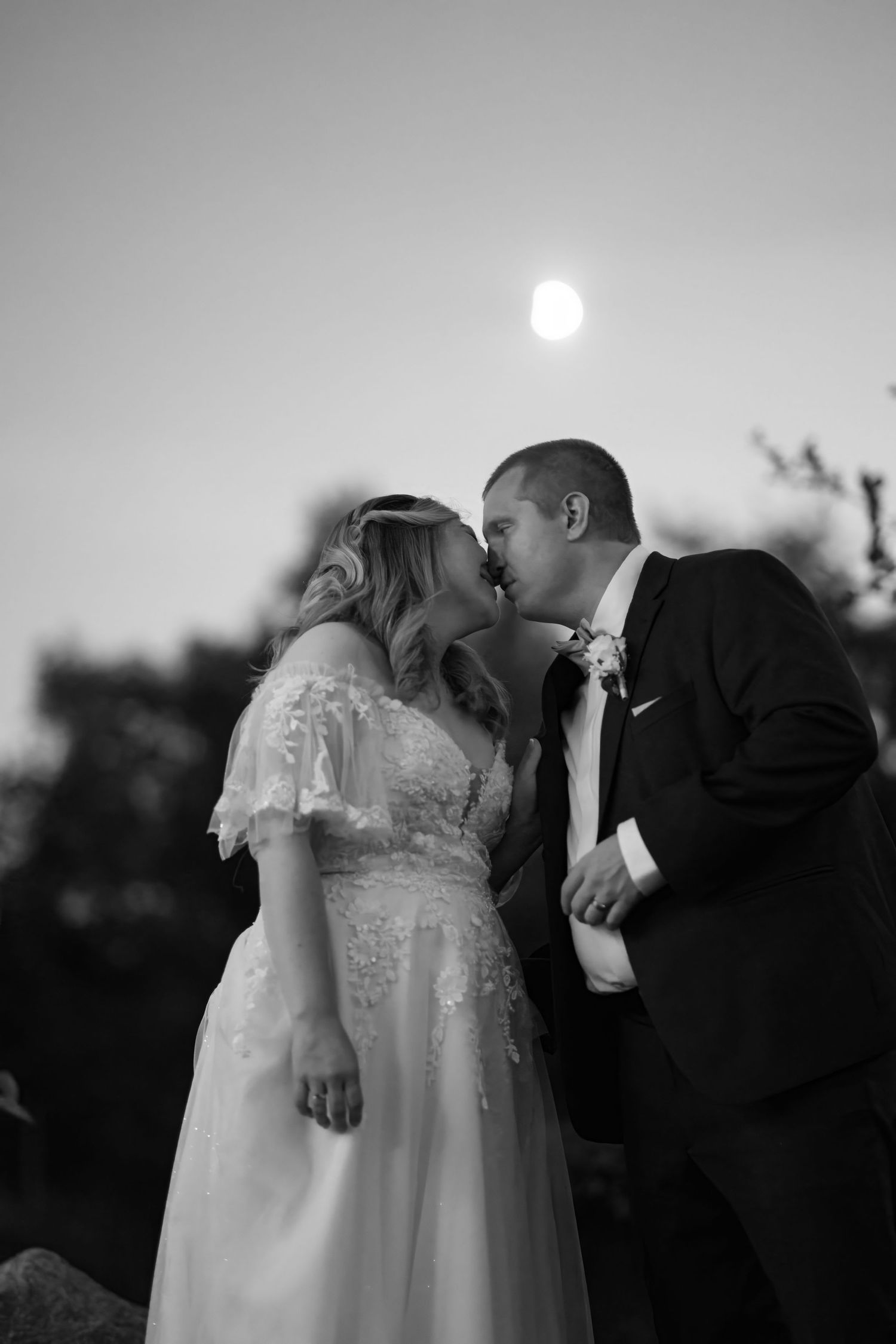 A romantic wedding couple shares a kiss at sunset in black and white.