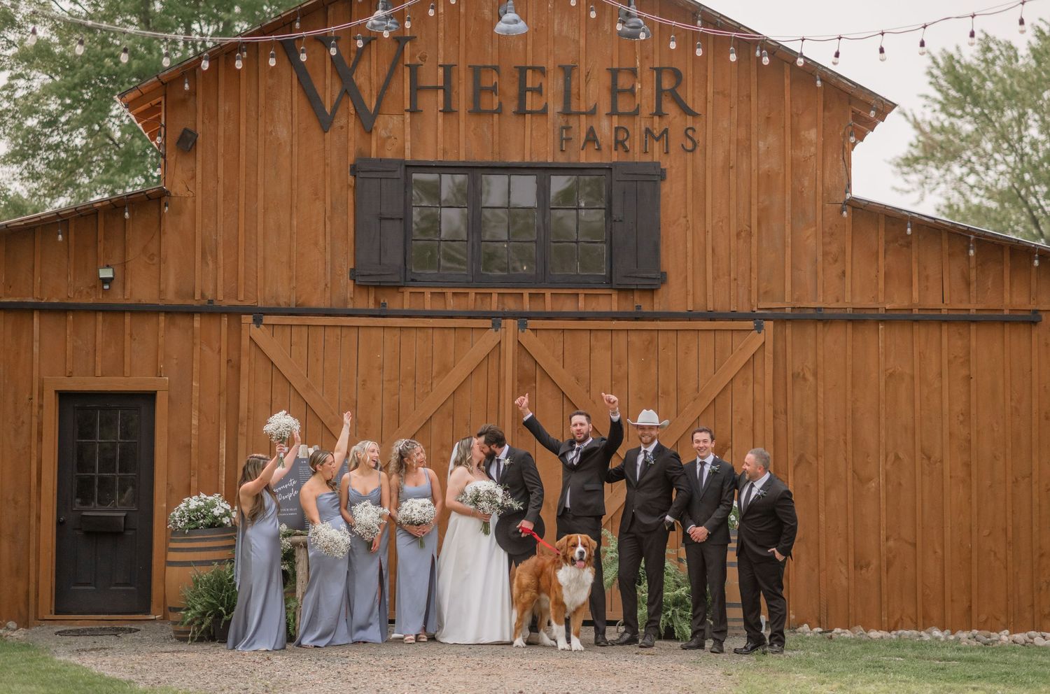 Wedding party poses in front of rustic wooden barn venue with string lights and elegant decor.