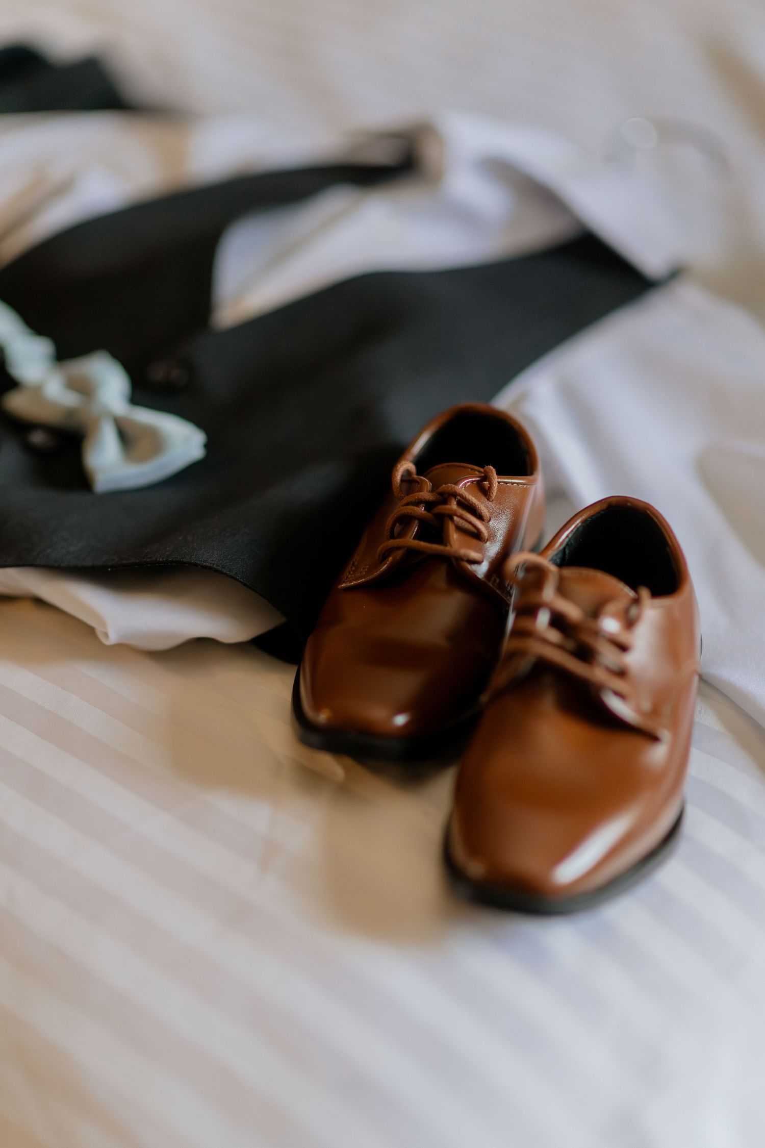 Brown leather dress shoes displayed on white bedding with a bow tie in the background.