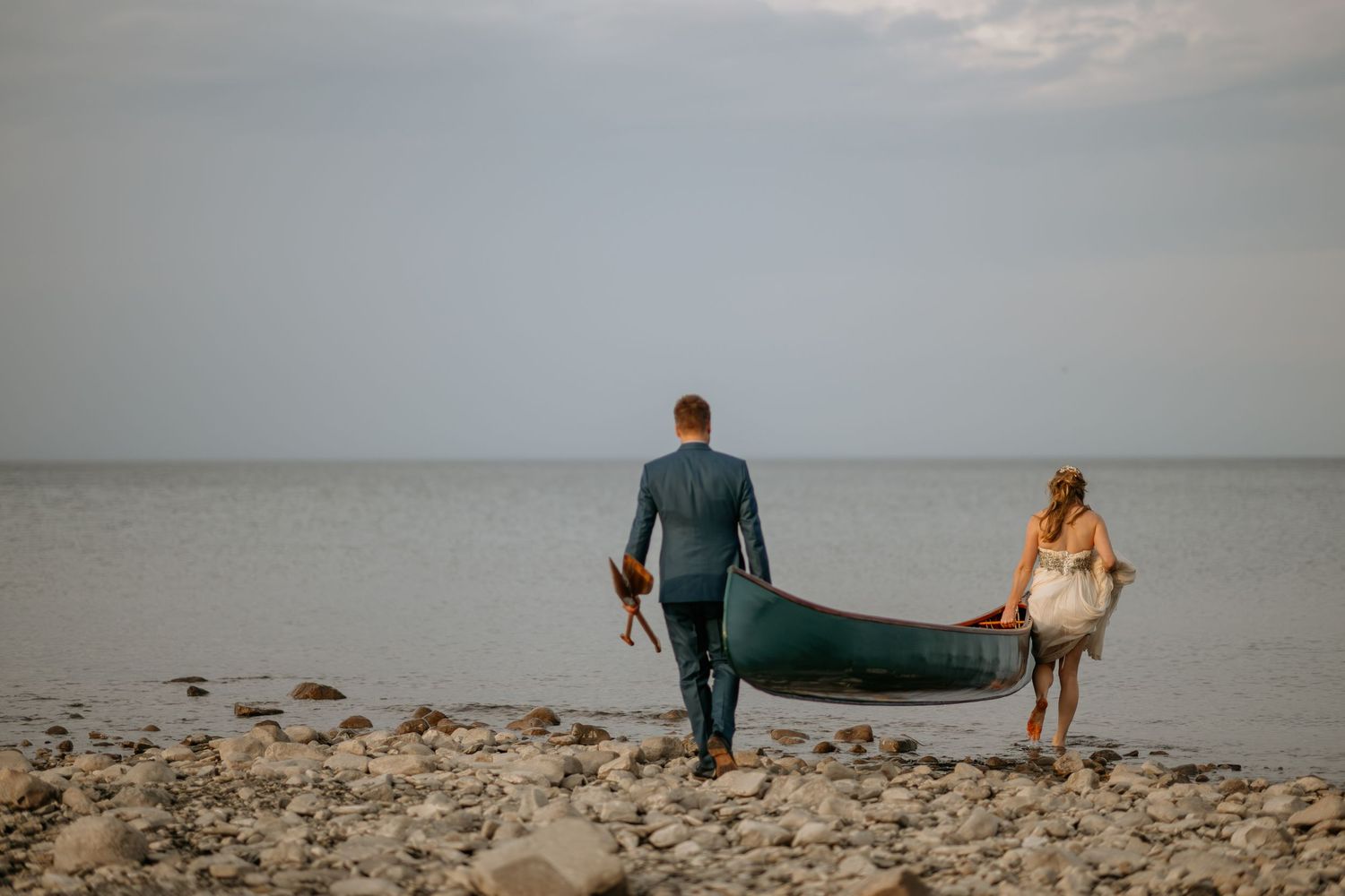 A couple carries a green canoe along a serene beach shoreline at dusk.