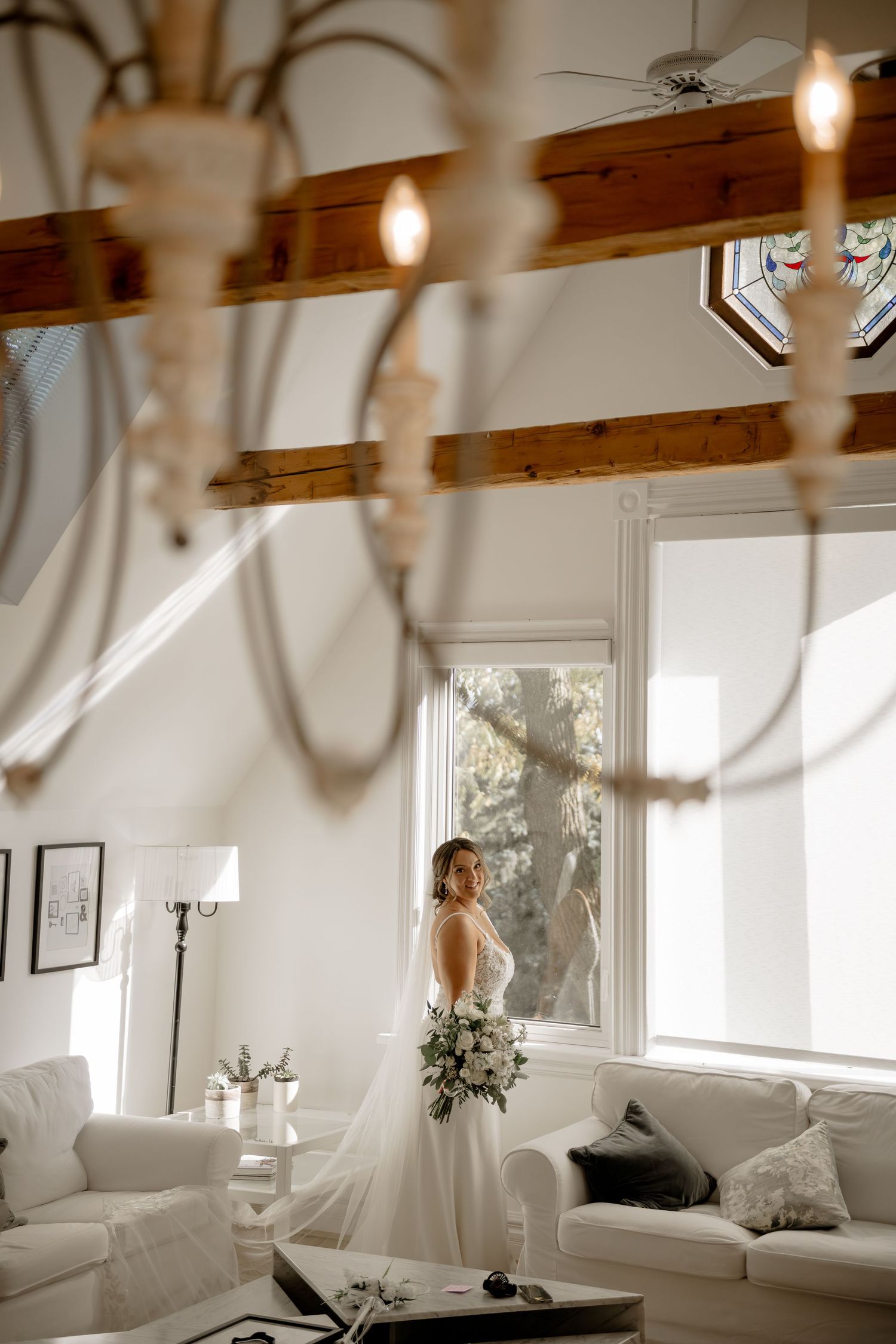 A figure in a white wedding dress stands by a bright window in a rustic room with exposed wooden beams and pendant lights.
