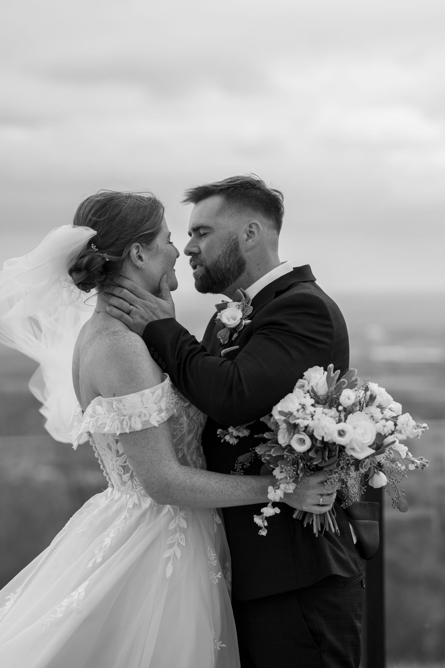 A romantic black and white wedding portrait of a couple embracing outdoors with a bouquet and cloudy sky backdrop.