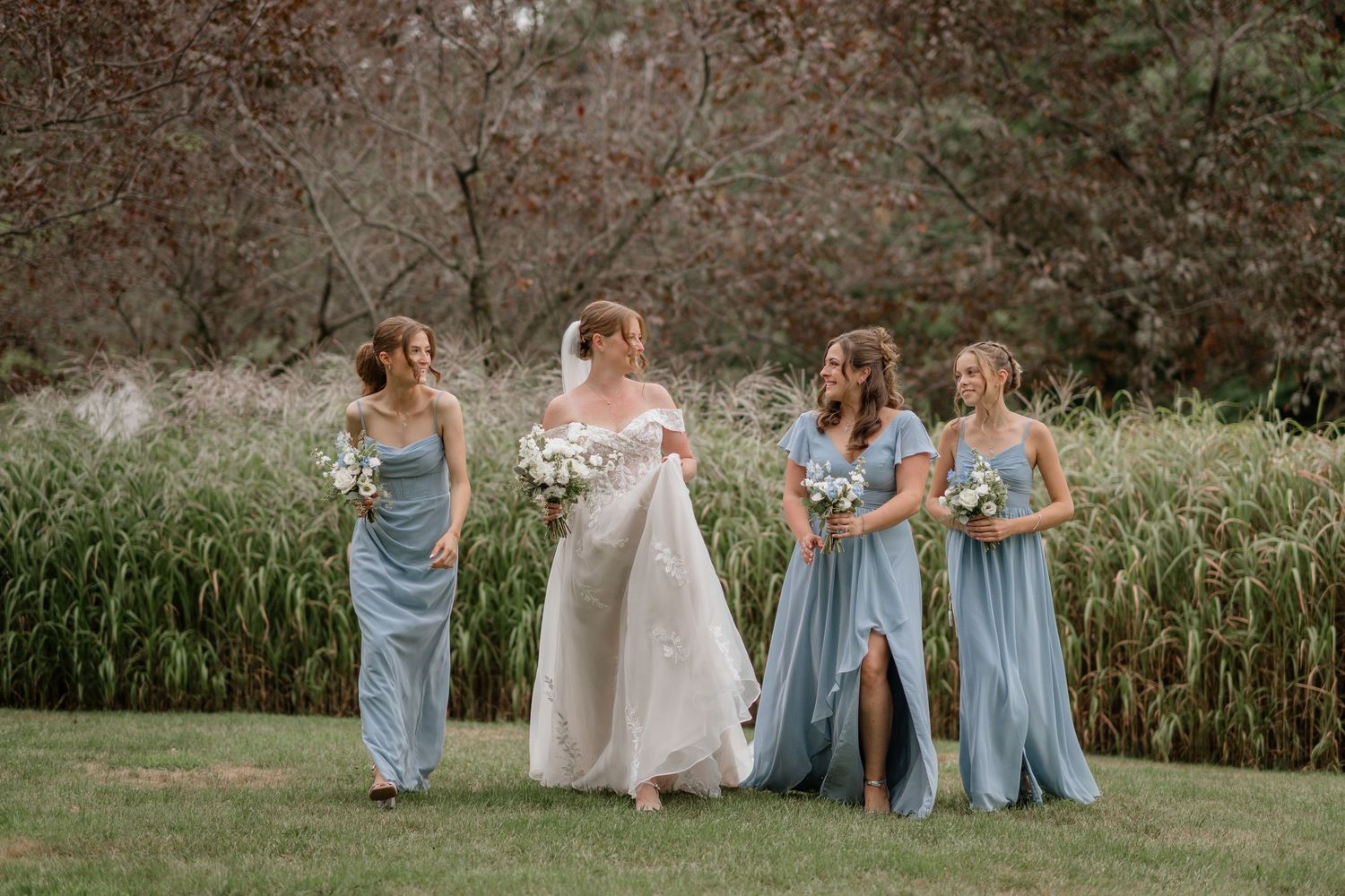 A group in light blue bridesmaid dresses and a white wedding gown walk together on a grassy lawn near tall reeds.