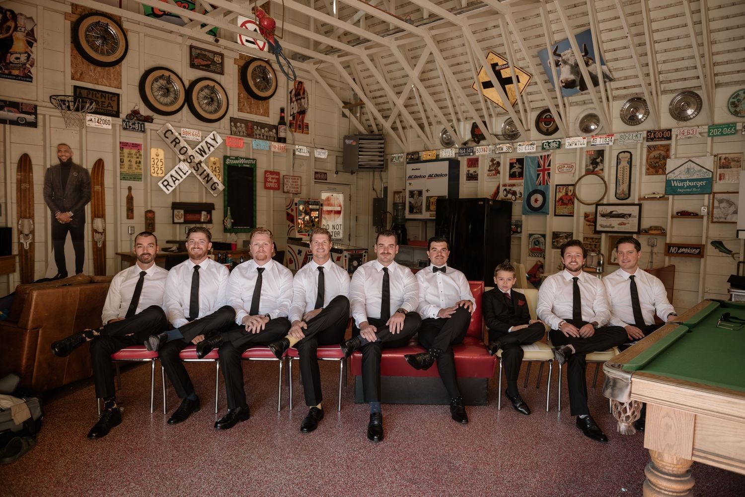 Groomsmen in black suits and white shirts sit on a red couch in a rustic venue decorated with vintage wall art.