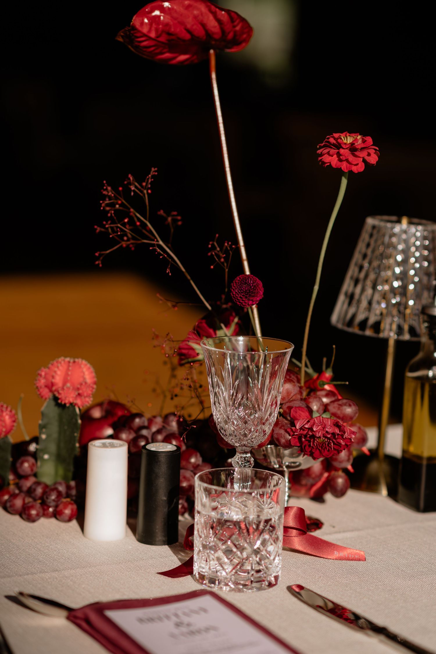 Elegant wedding table decor featuring red flowers, crystal glasses, and grapes against a dark background.