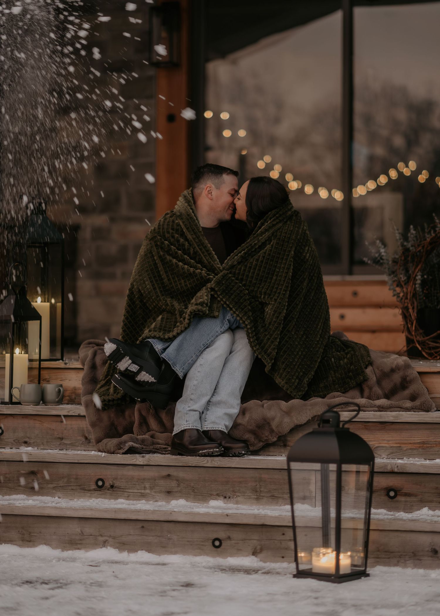 A couple sits together on a snowy bench at night with string lights glowing in the background.