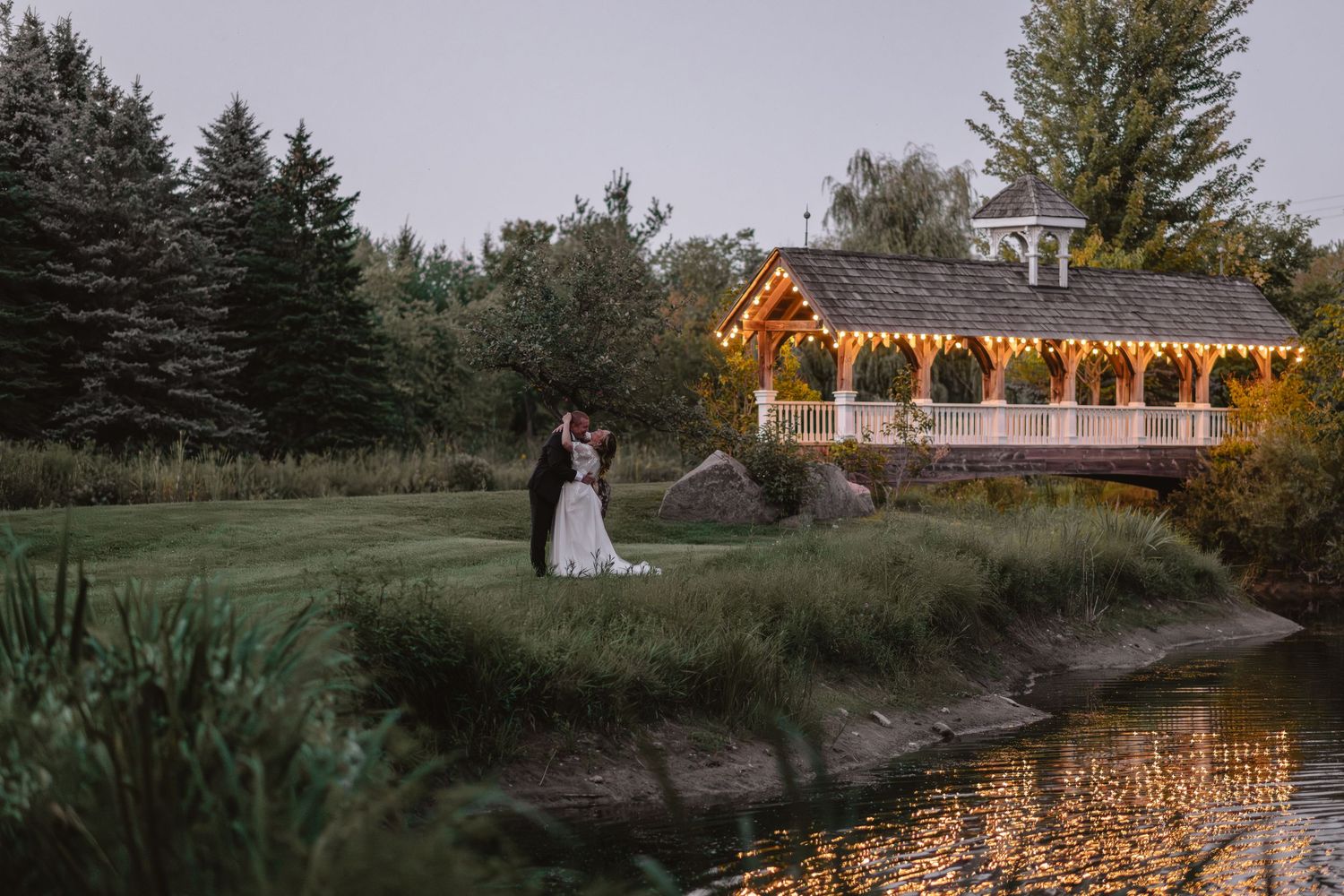 A romantic evening scene at a gazebo decorated with string lights reflecting in a pond at dusk.