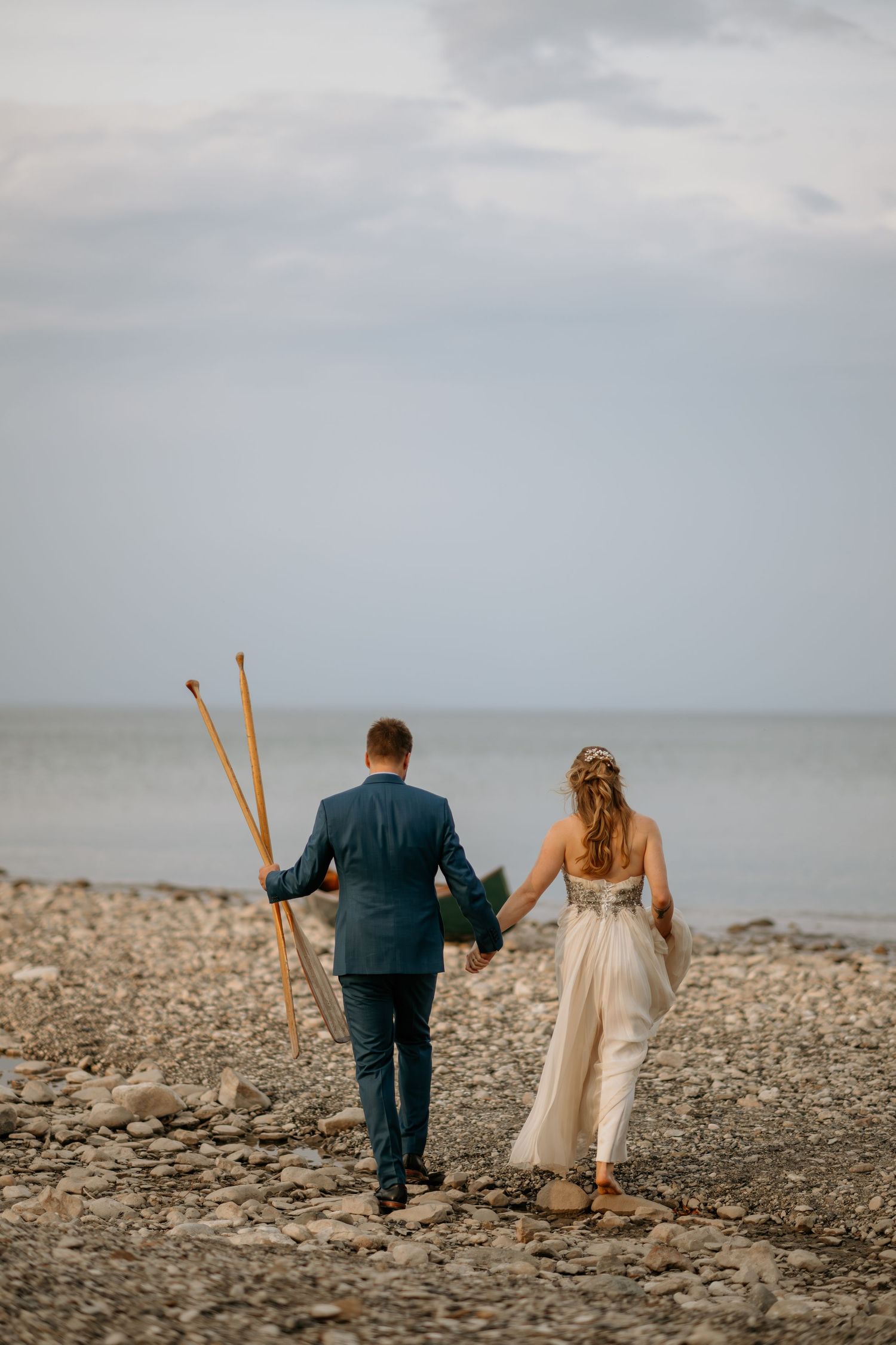 A couple walks along a rocky beach shore carrying tall wooden staffs at sunset.