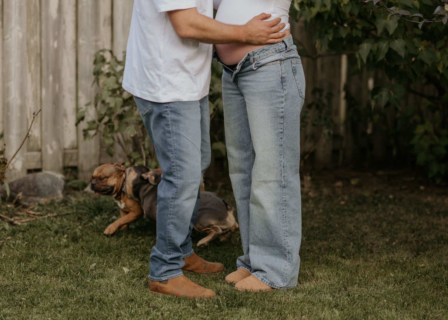 Casual couple with dog poses in backyard wearing jeans and white shirts.