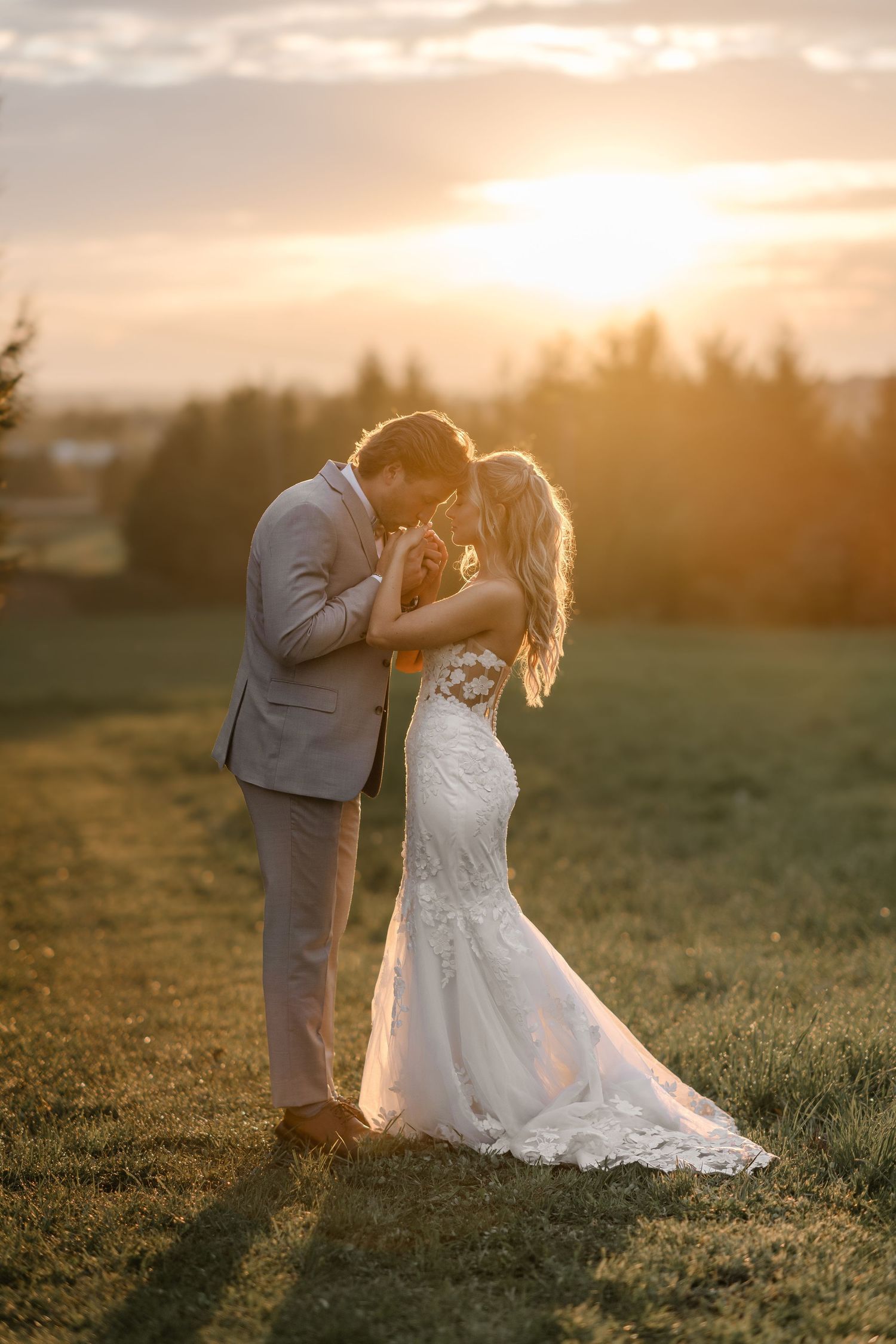 Bride and groom share a romantic moment at golden sunset in a green field, bride in lace mermaid gown.