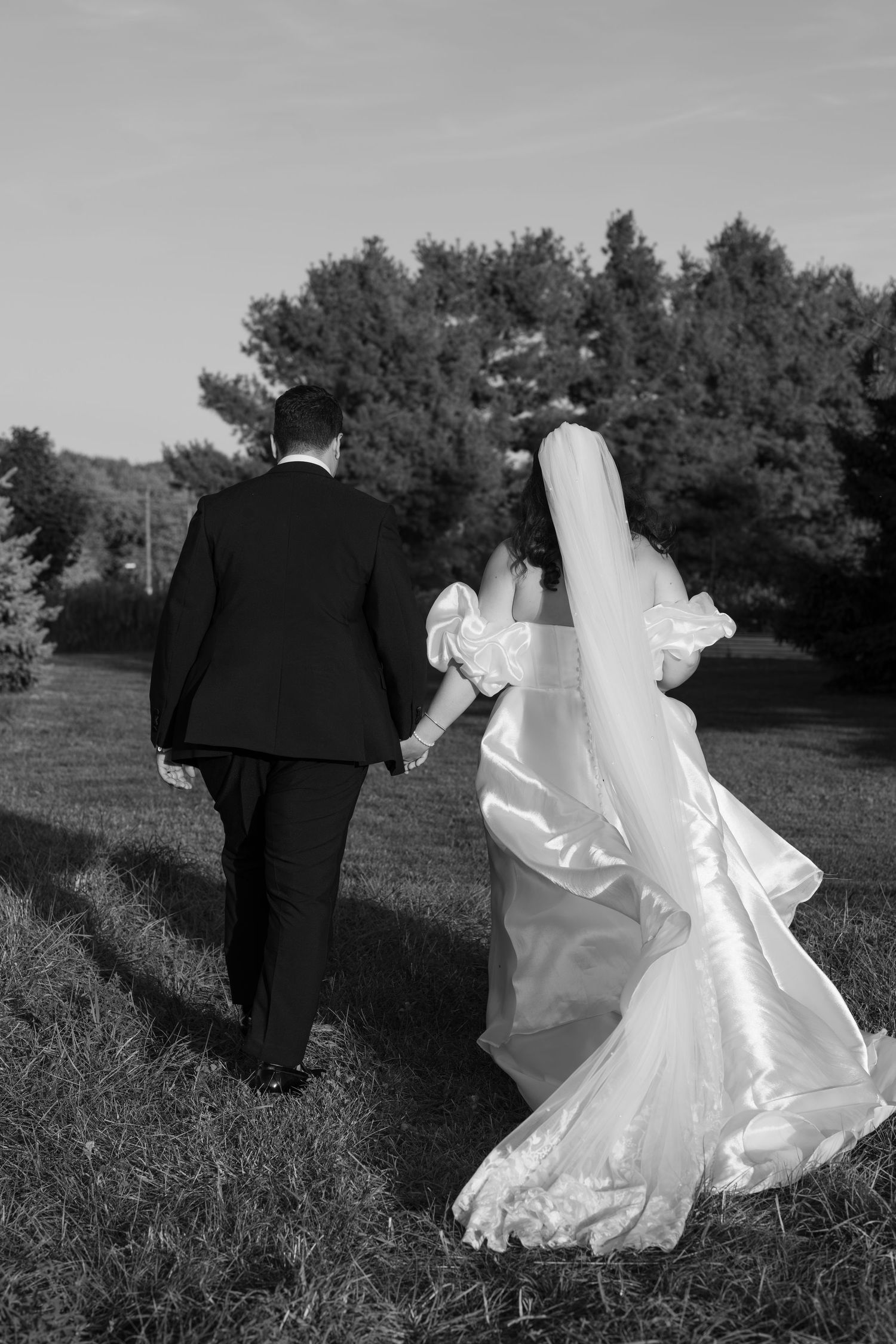 A black and white photo of newlyweds walking hand in hand through a grassy field.