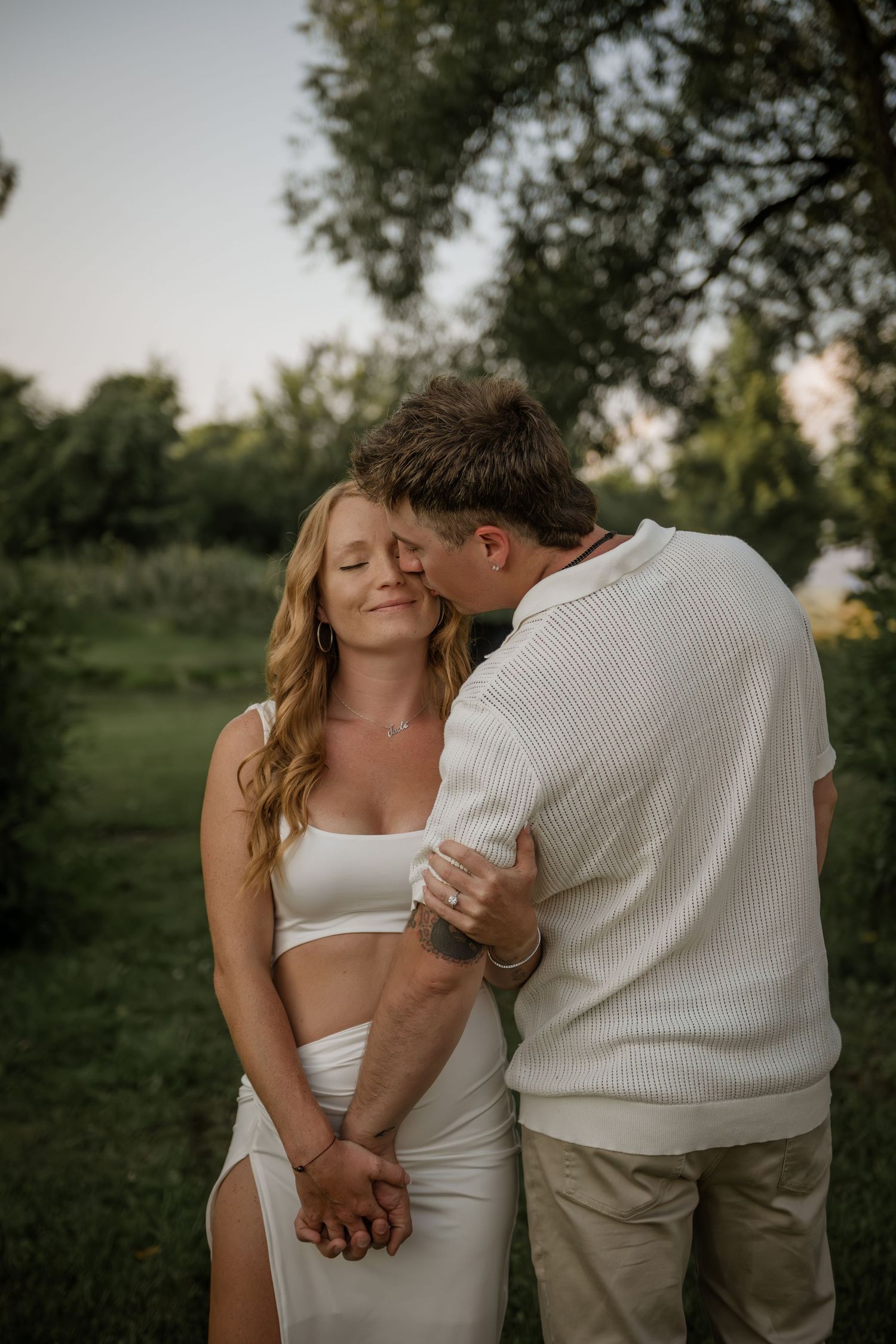 Intimate couple embraces in casual white outfits against natural outdoor setting.