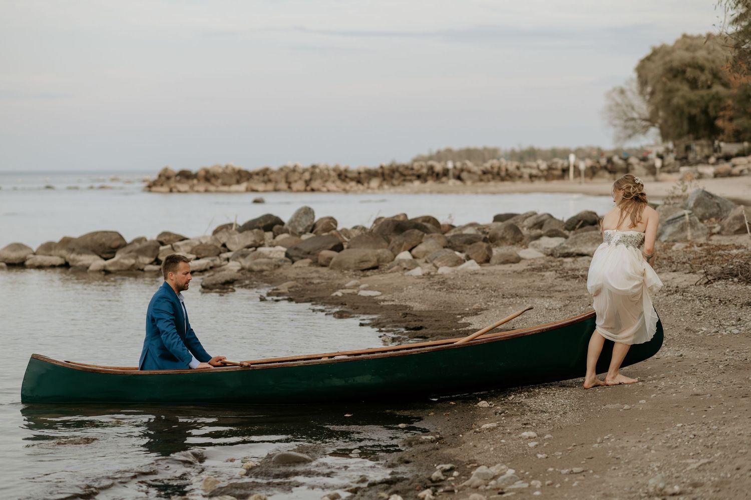 A green canoe rests on a rocky beach shore with calm waters and distant coastline in view.
