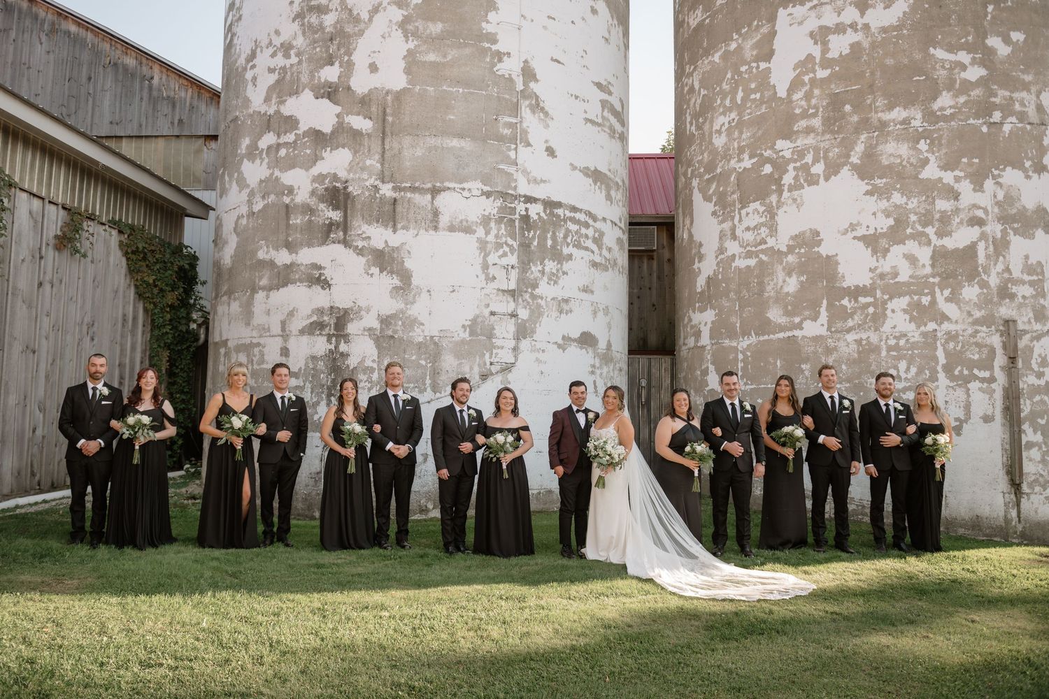A wedding party in black dresses poses in front of rustic concrete silos on a grassy lawn.