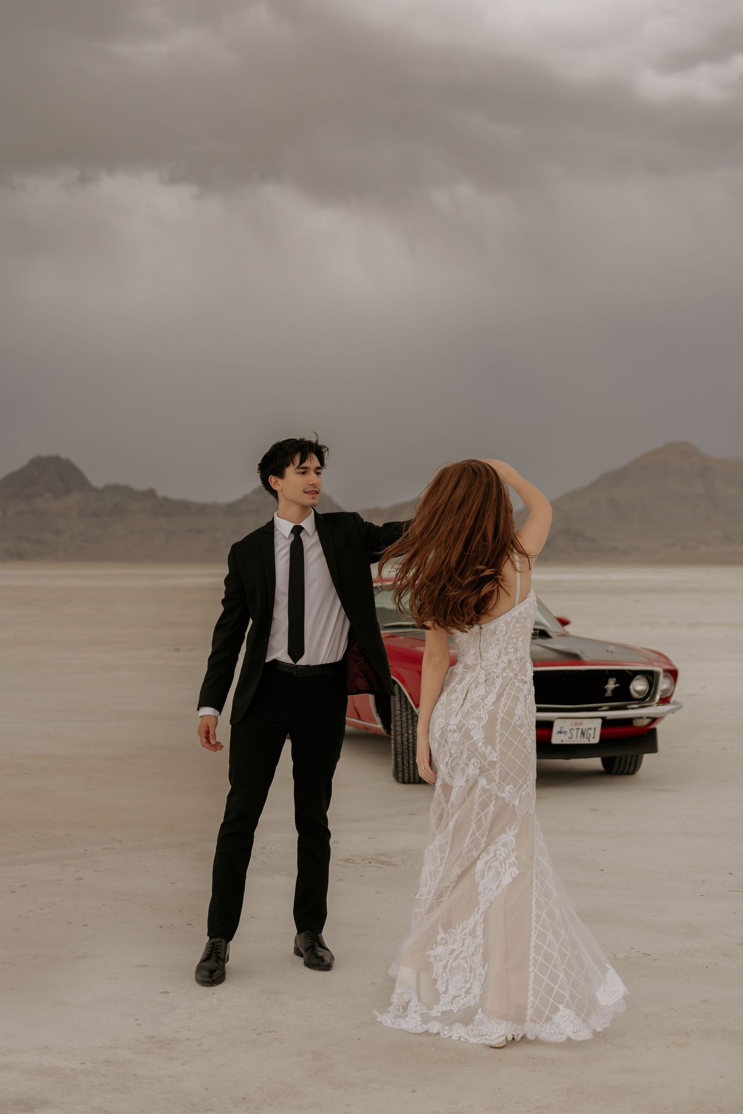 A couple in formal wedding attire dances together on a scenic salt flat with a vintage car in the background.