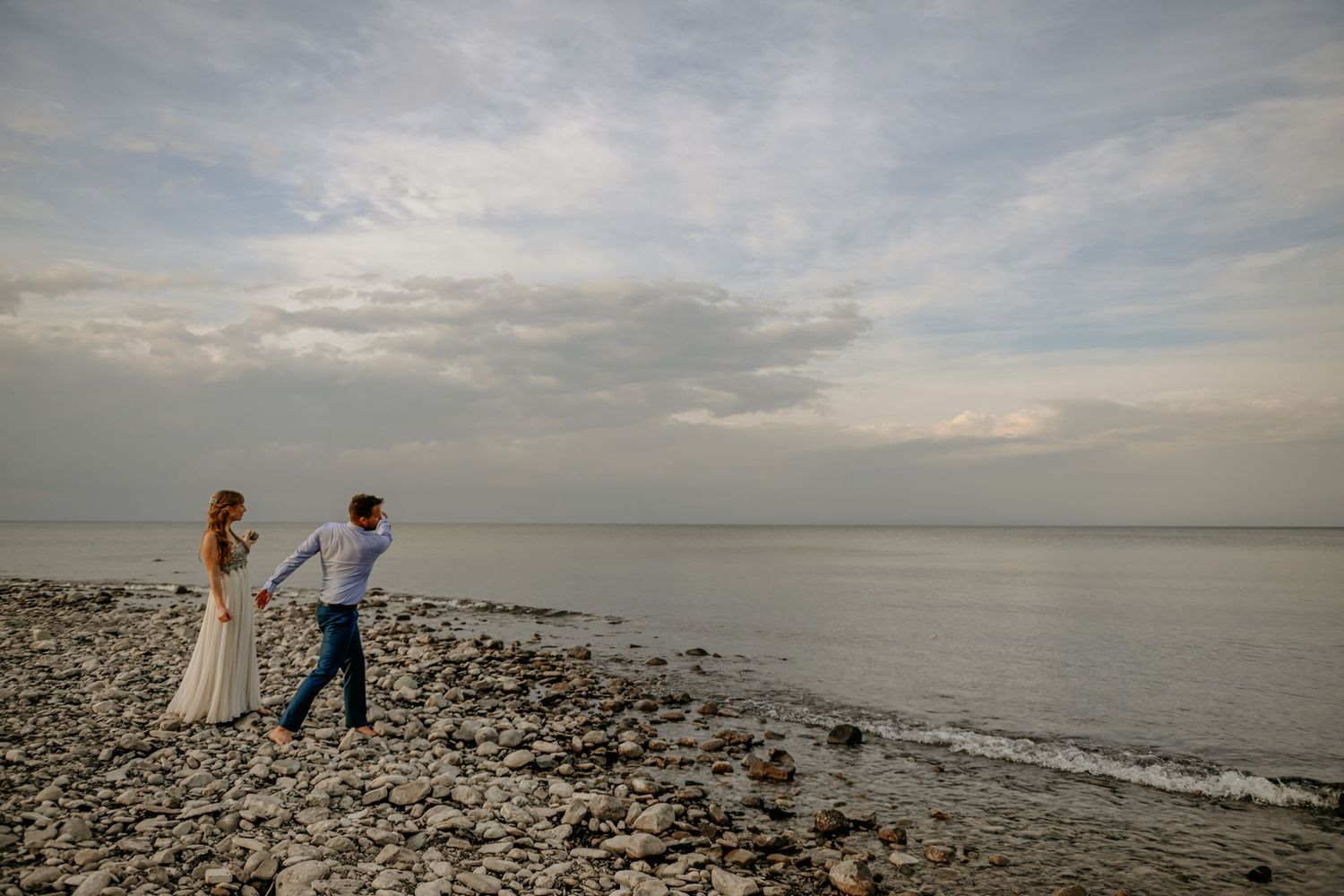 Wedding couple standing on rocky shoreline during golden hour with dramatic cloudy sky.