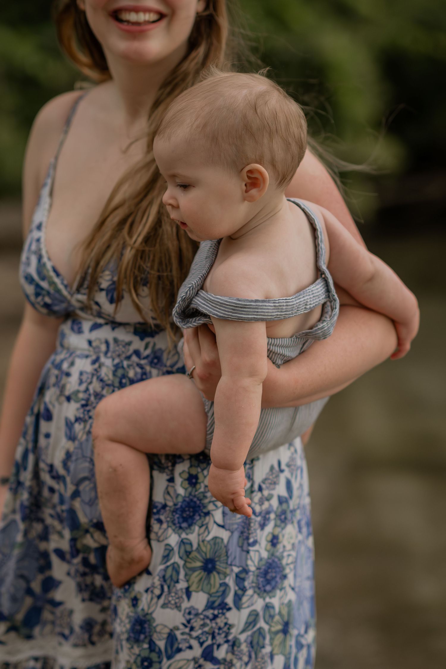 Woman in floral dress holding baby wearing striped romper against natural backdrop.