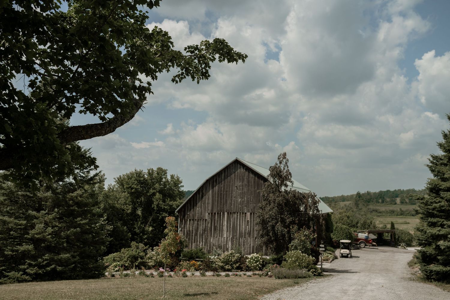 A rustic wooden barn sits among trees and landscaping on a winding gravel driveway under cloudy skies.