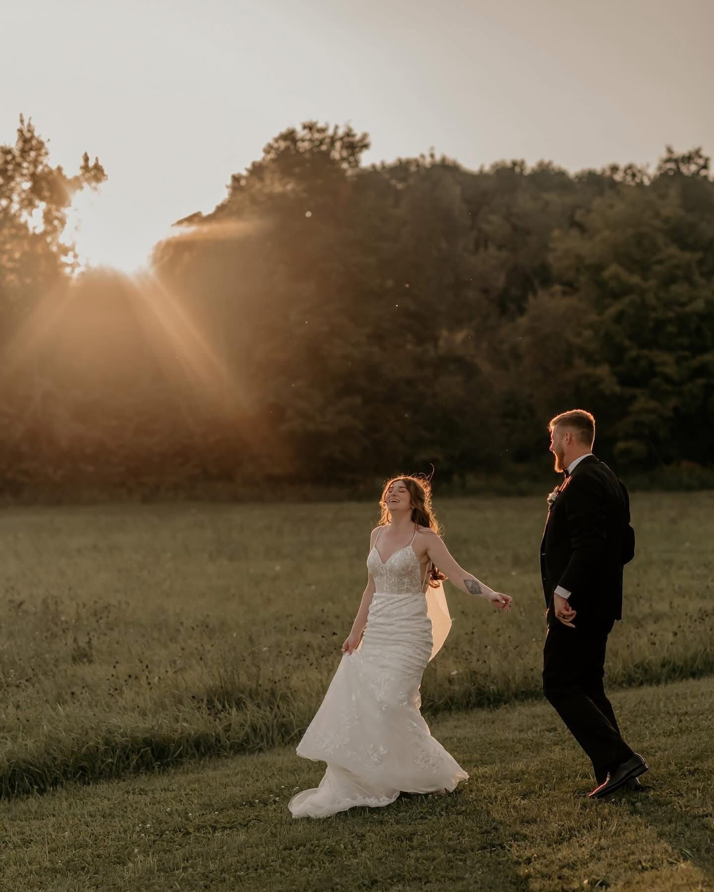 A couple in wedding attire dances playfully in a sunlit meadow during golden hour.