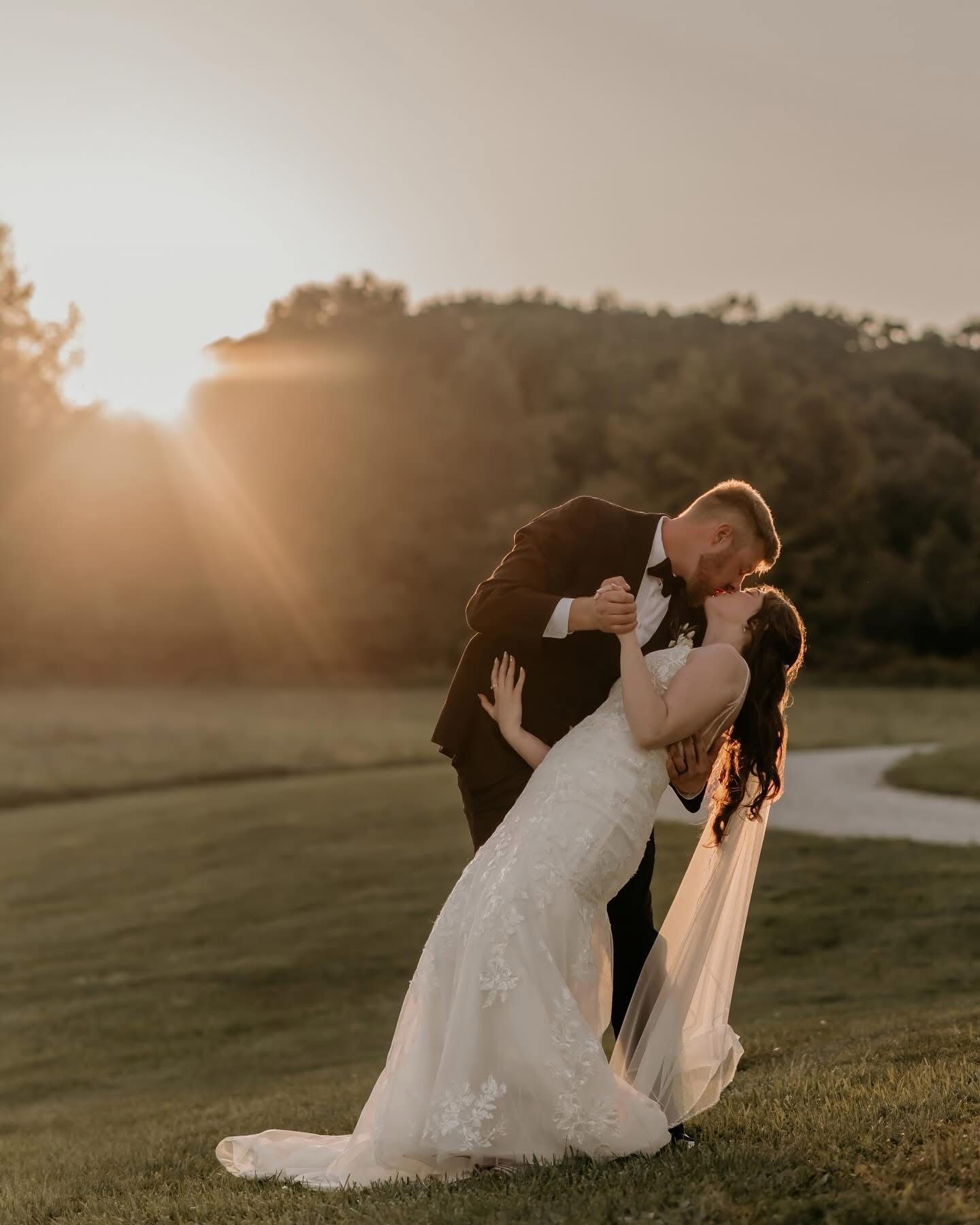 Romantic wedding couple embraces in a dramatic dip pose during golden hour sunset on a lush green hillside.