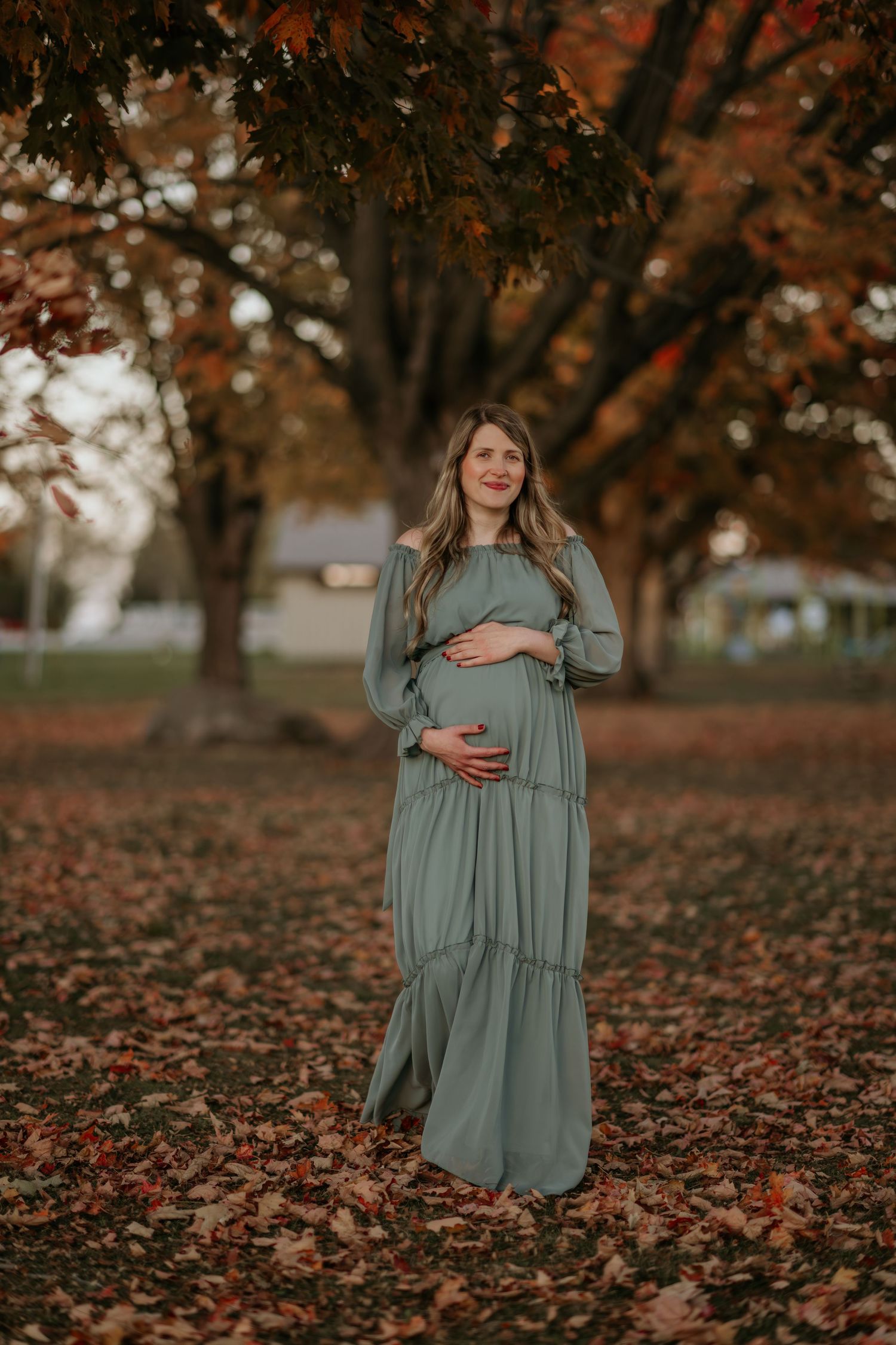 Autumn maternity photos with fallen leaves and trees showing woman in sage green dress.