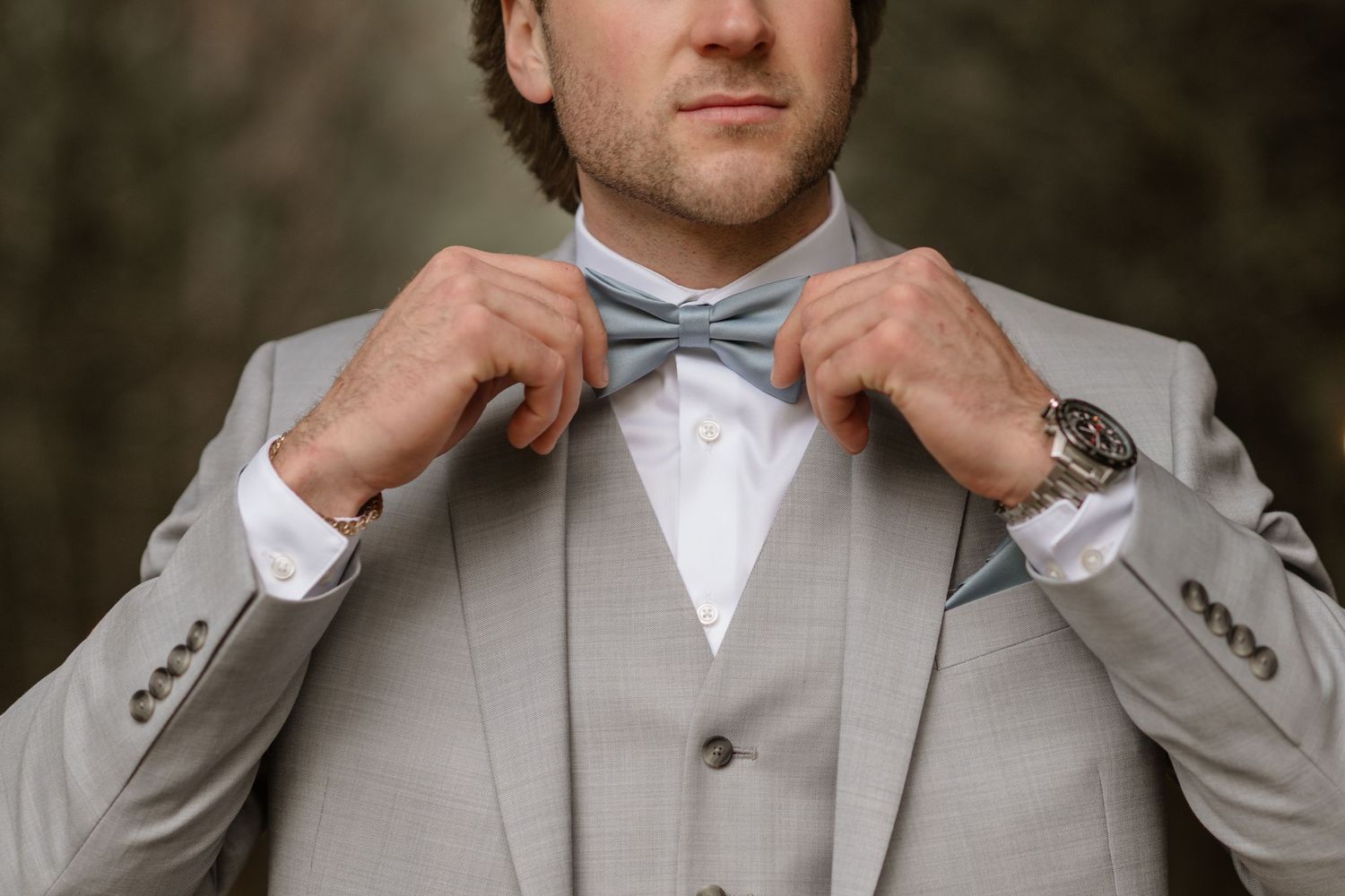 Man in light gray suit adjusting blue bow tie, wearing a watch, elegant wedding groom attire.