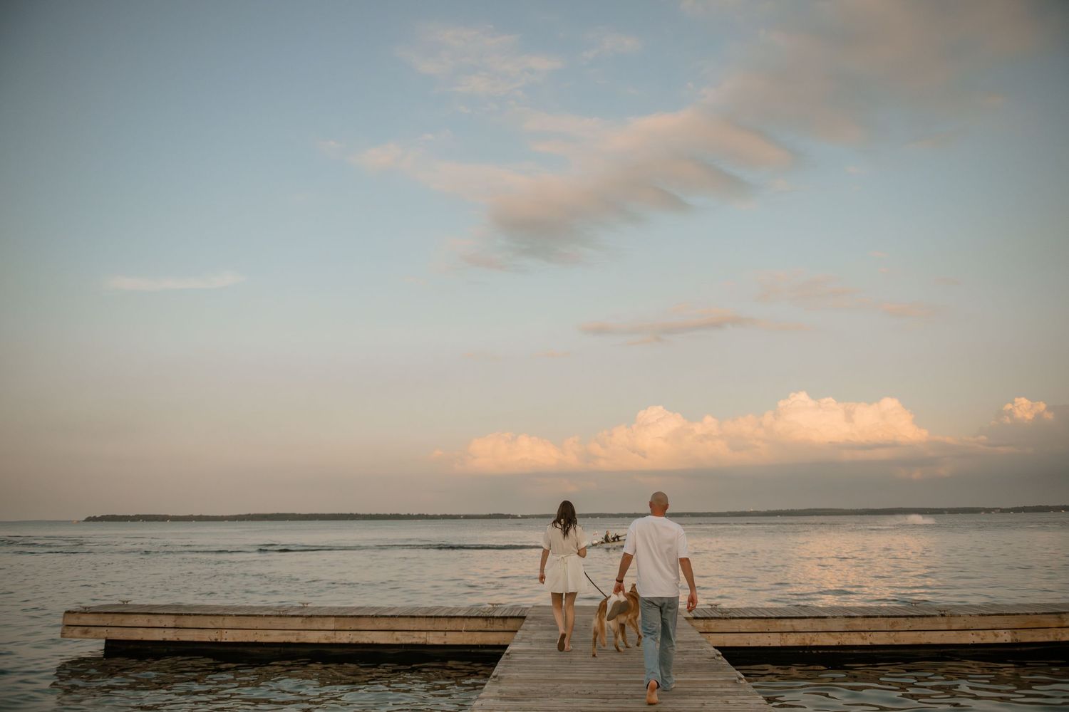 Two people walking hand in hand on a wooden pier at sunset with pink clouds reflecting on calm ocean waters.