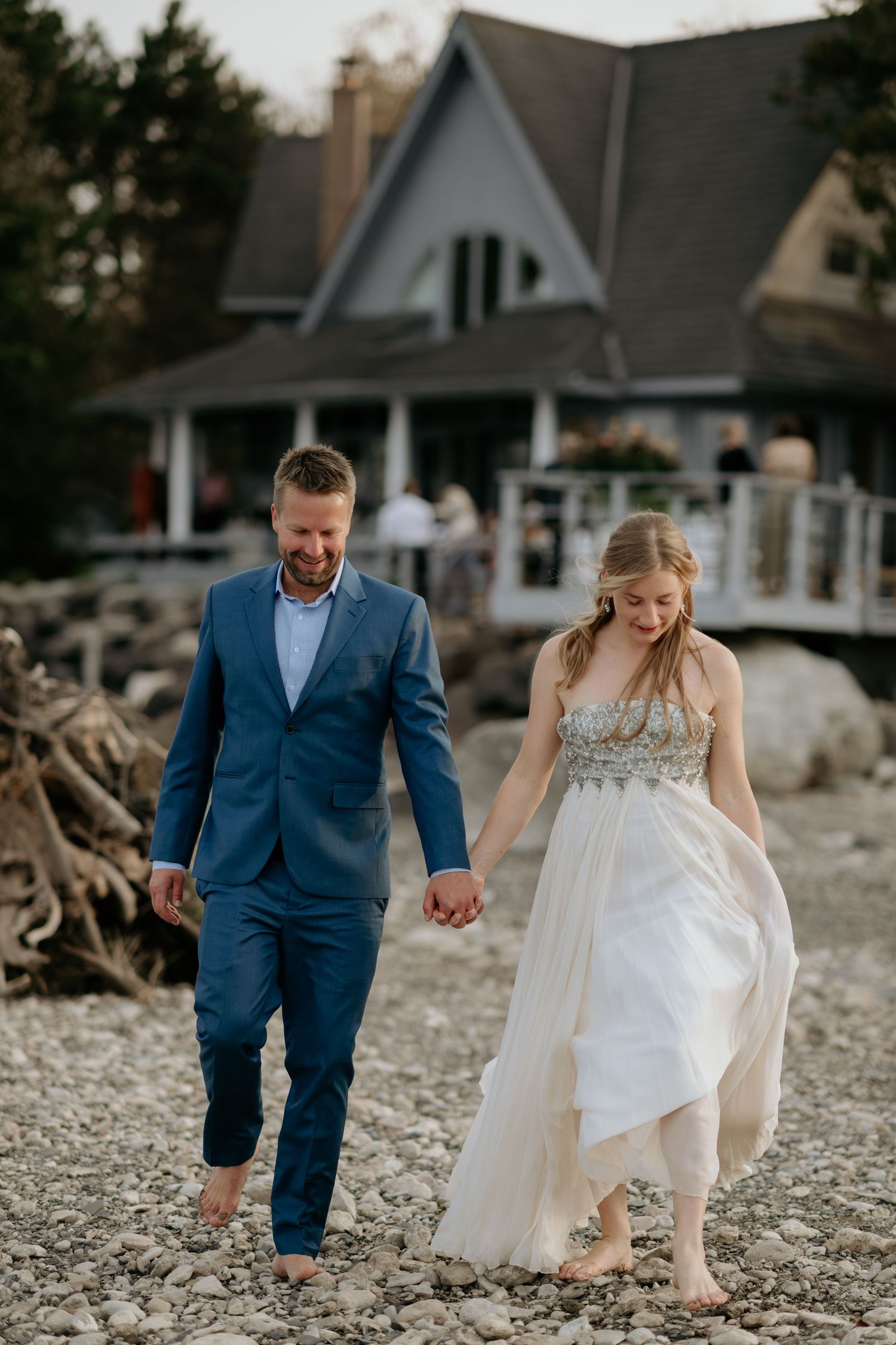 A couple walks hand in hand along a rocky beach shoreline with coastal houses in the background.