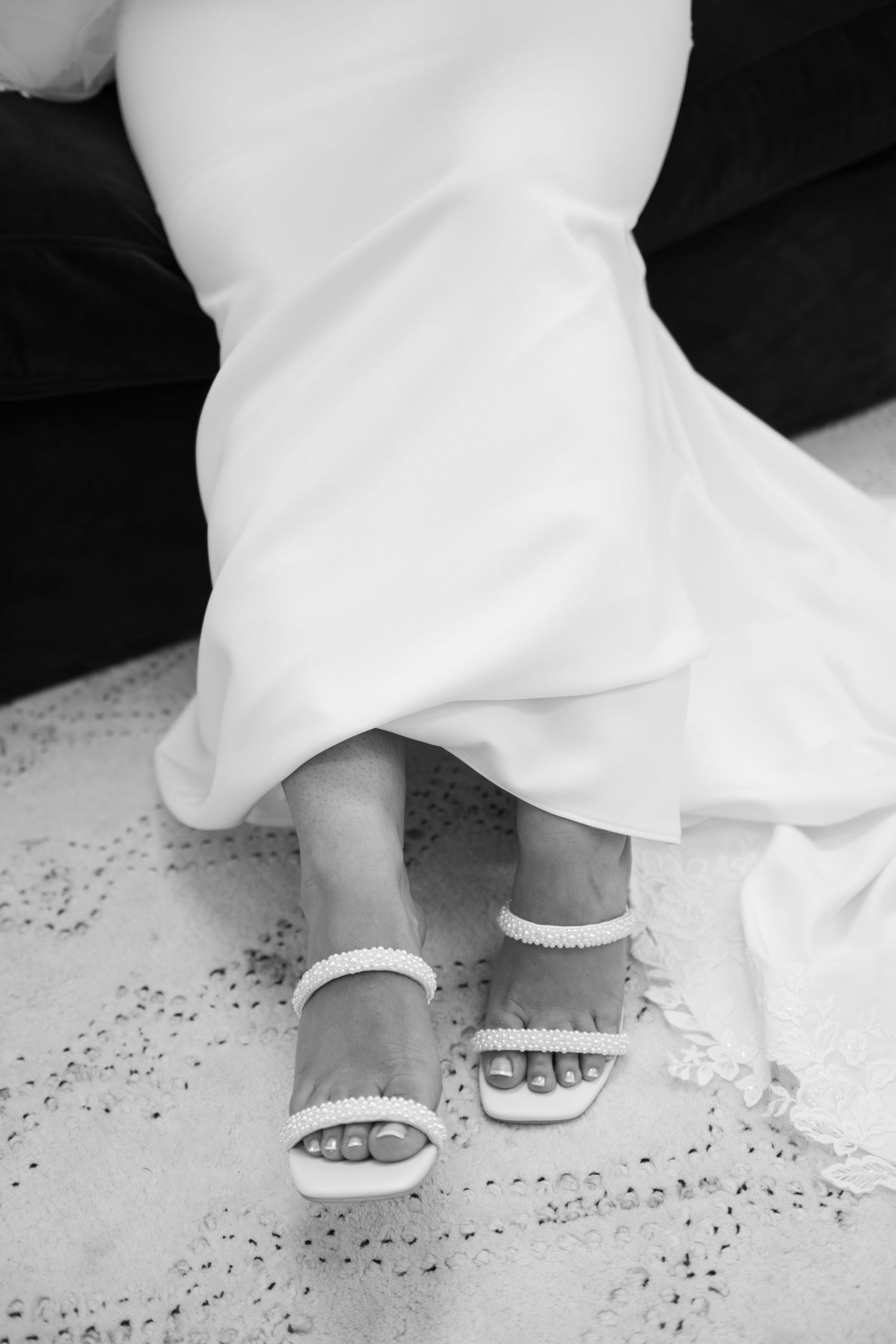 Close-up view of elegant bridal sandals peeking out from under a flowing white wedding dress on a speckled floor.