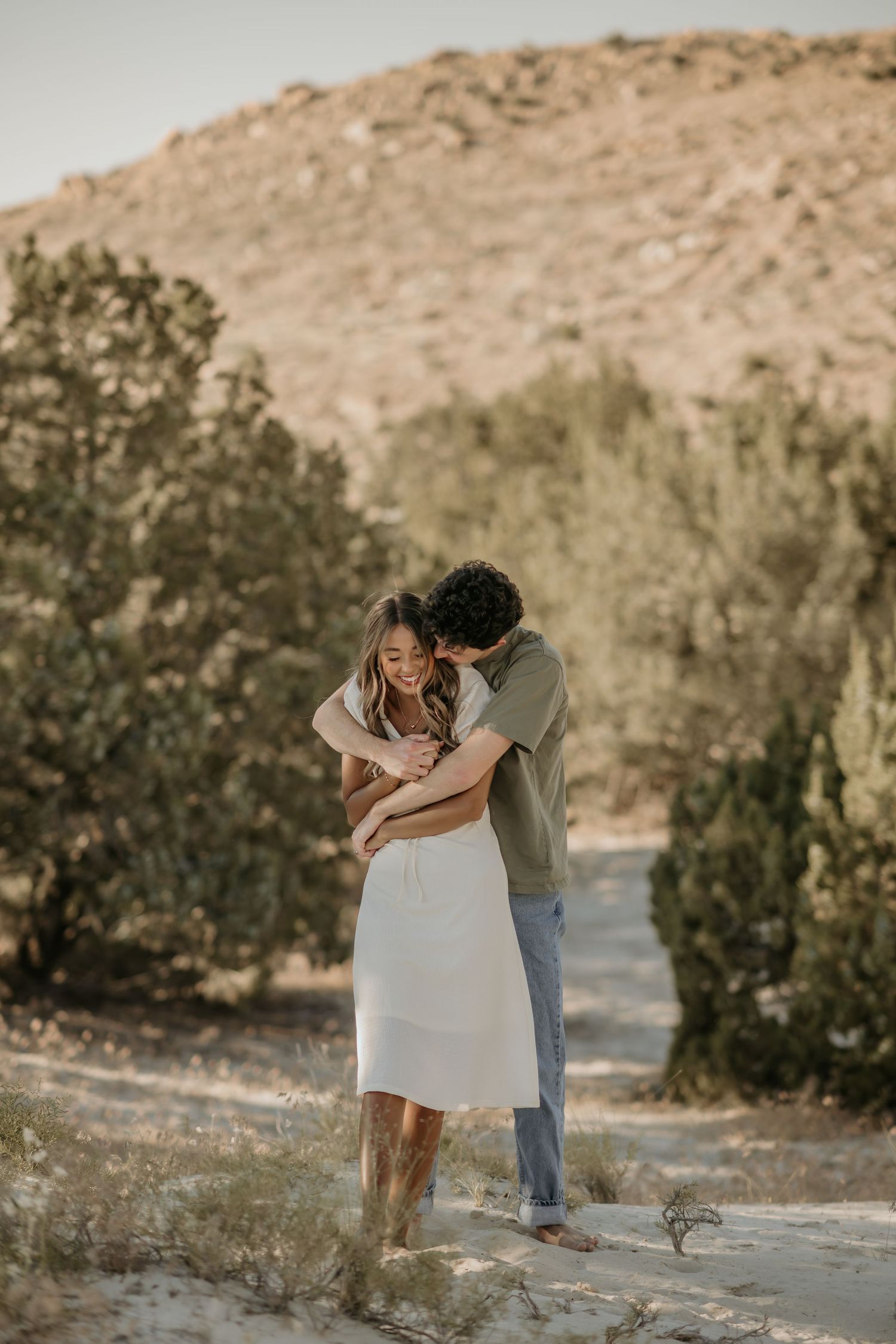 A couple embraces on a desert trail with brown hills and shrubs in the background during golden hour.