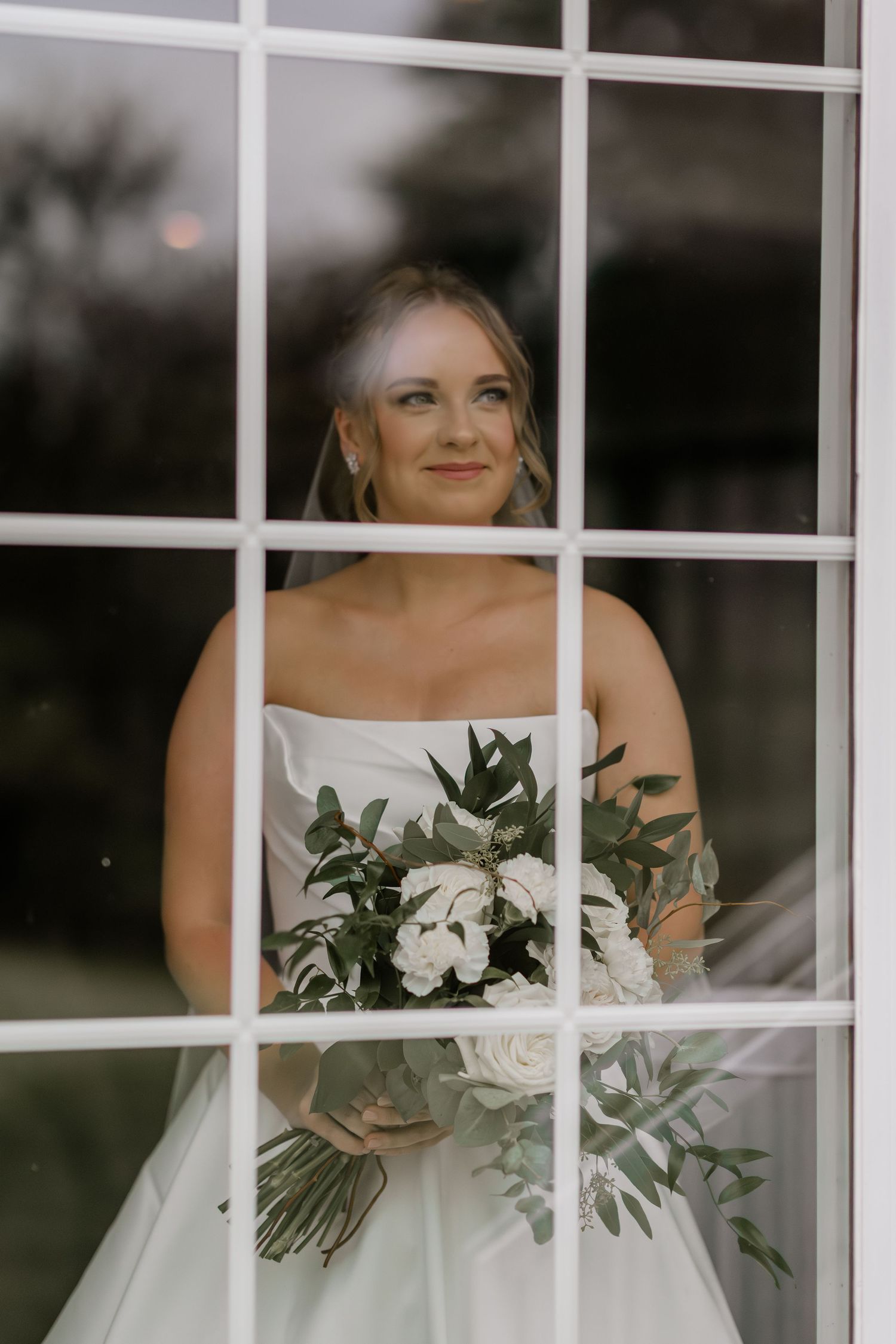 White wedding dress and bouquet framed through elegant window panes in dramatic lighting.