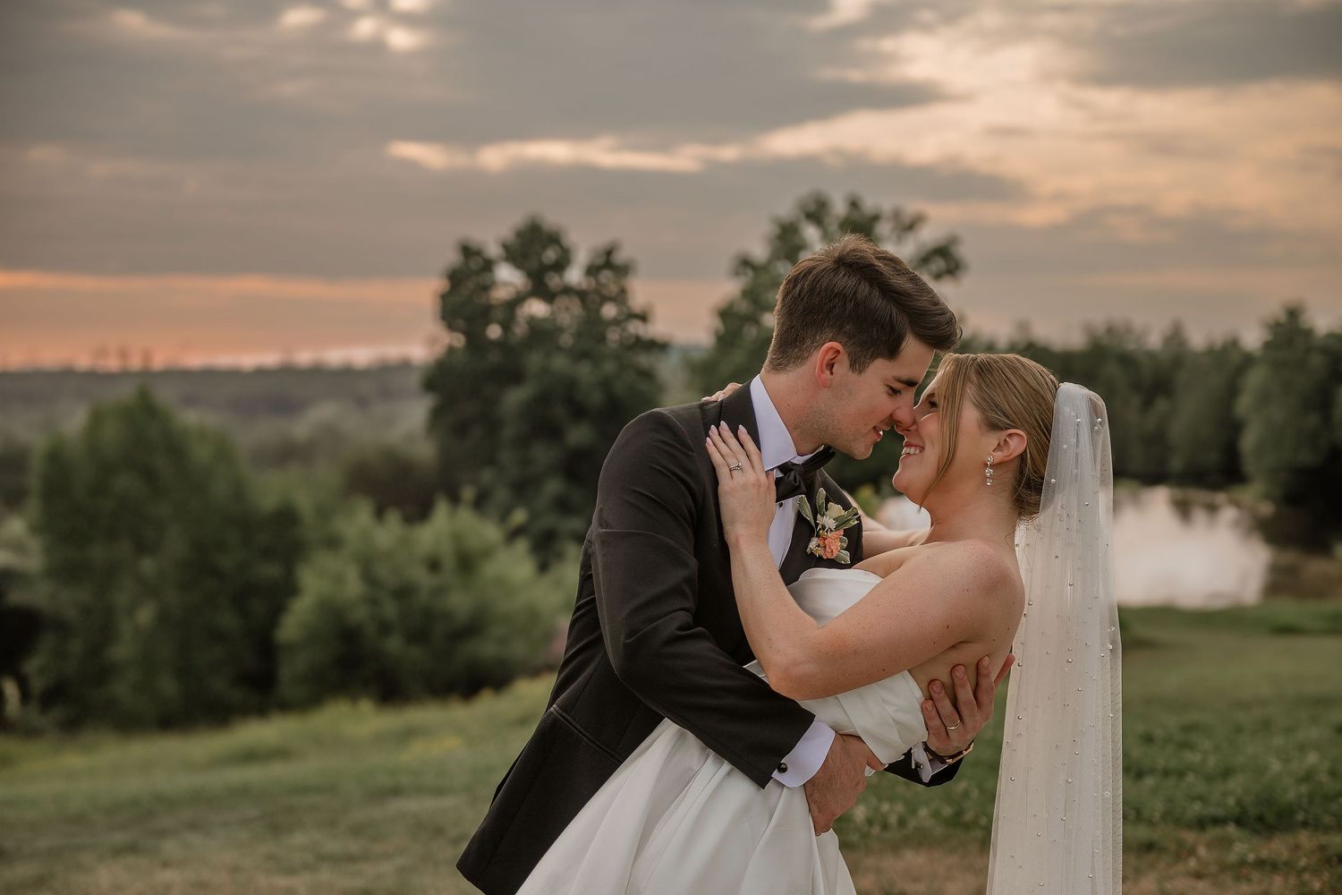 Newlywed couple shares romantic kisses at sunset in a scenic outdoor setting with trees and golden light.