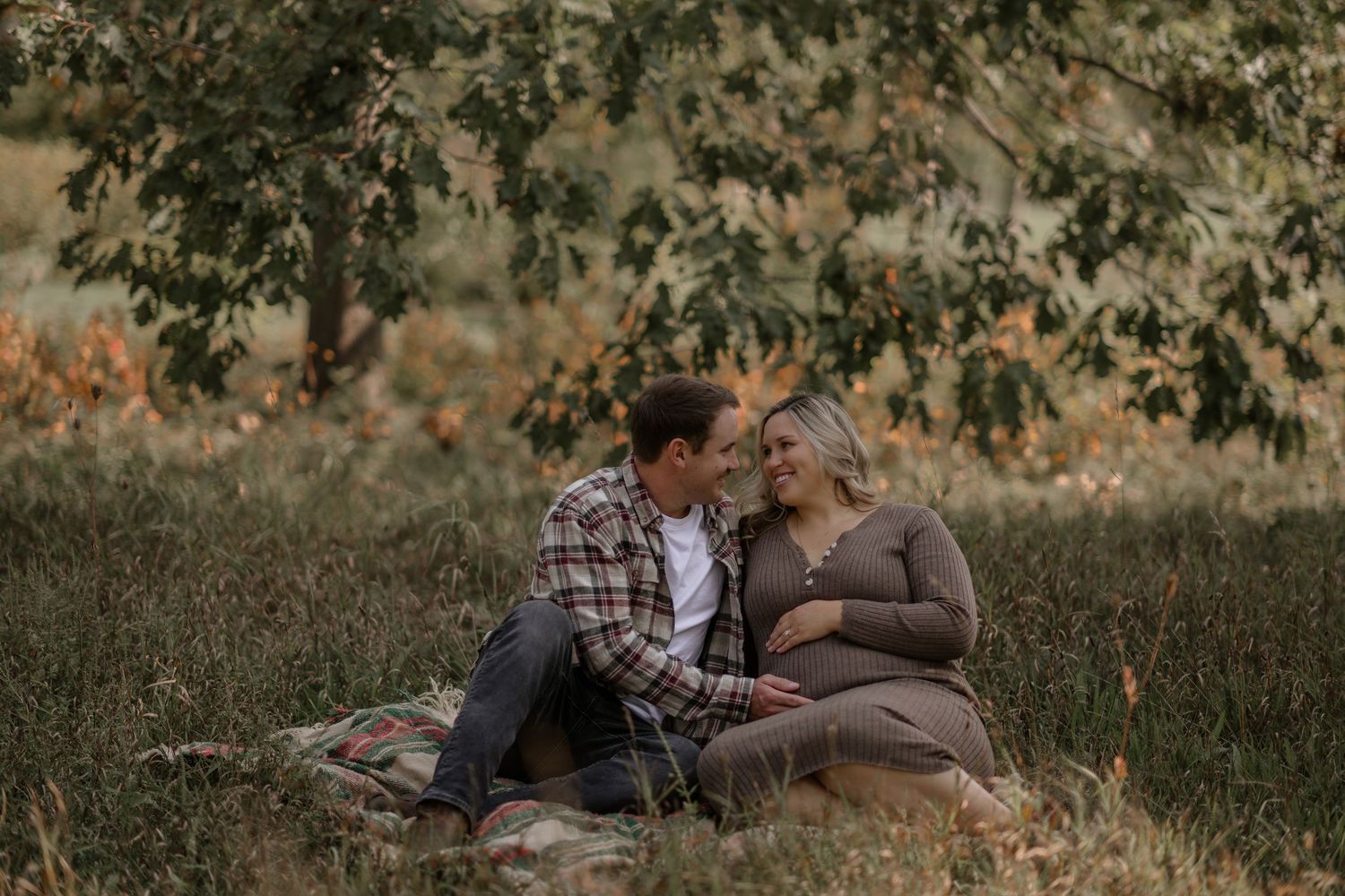 Couple sits together on grass during autumn maternity photoshoot.