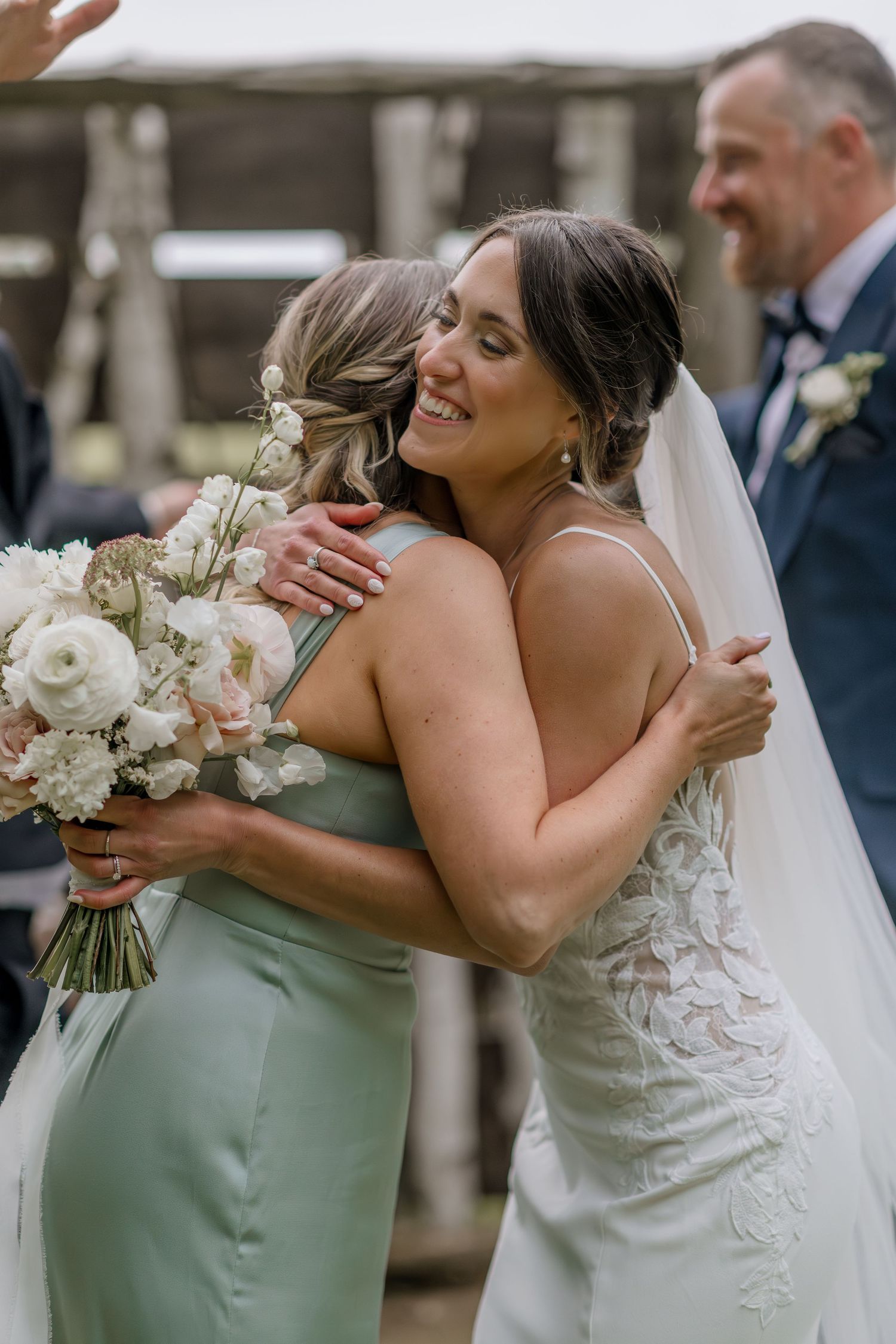 Bride in lace dress embraces bridesmaid wearing sage green dress while holding white bouquet.