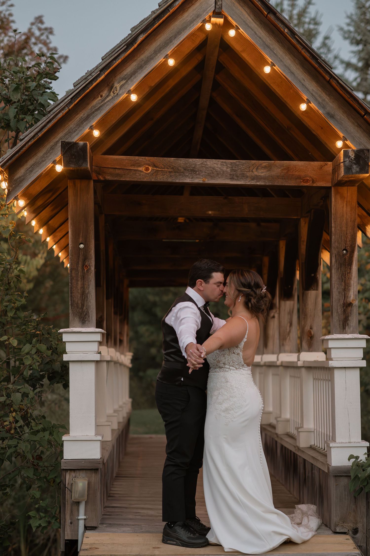 A couple shares a romantic dance under a rustic wooden pavilion decorated with string lights at dusk.