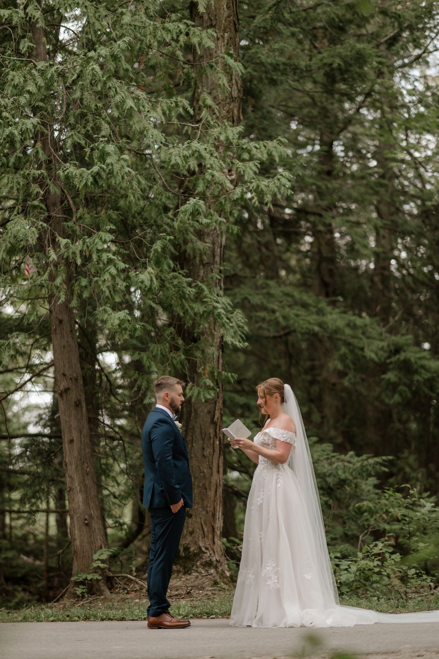 A wedding ceremony takes place on a path surrounded by tall evergreen trees in a forest setting.