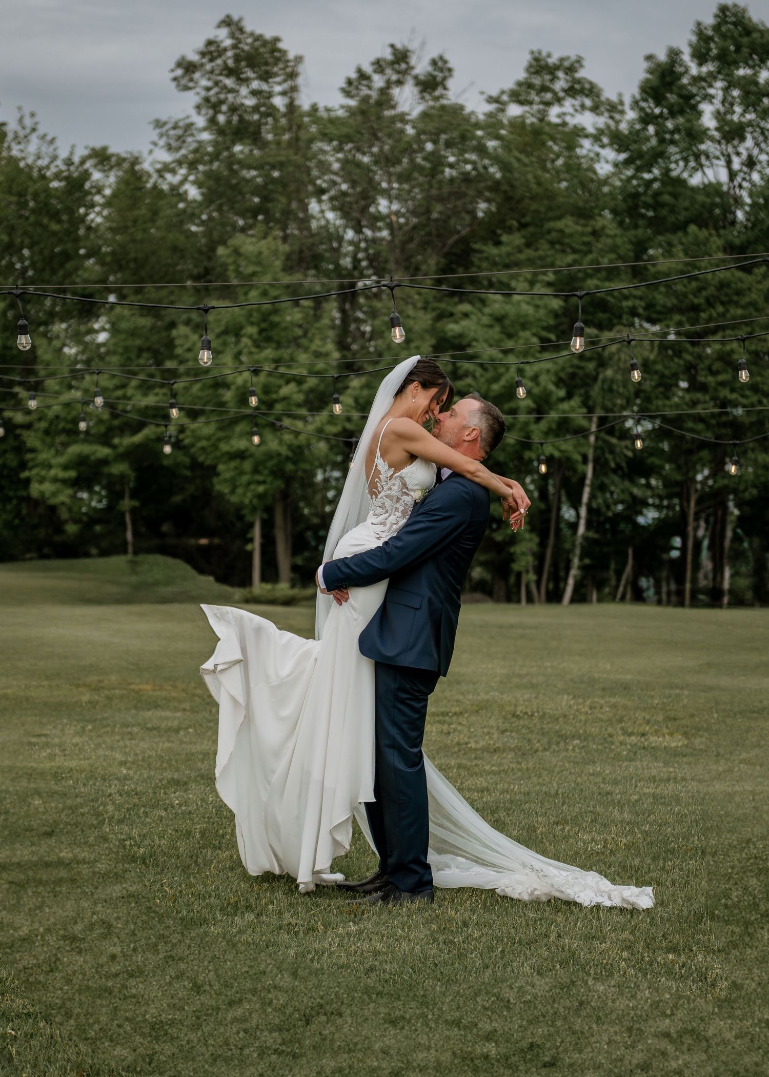 A romantic wedding couple dips and dances on a grassy lawn with string lights overhead.