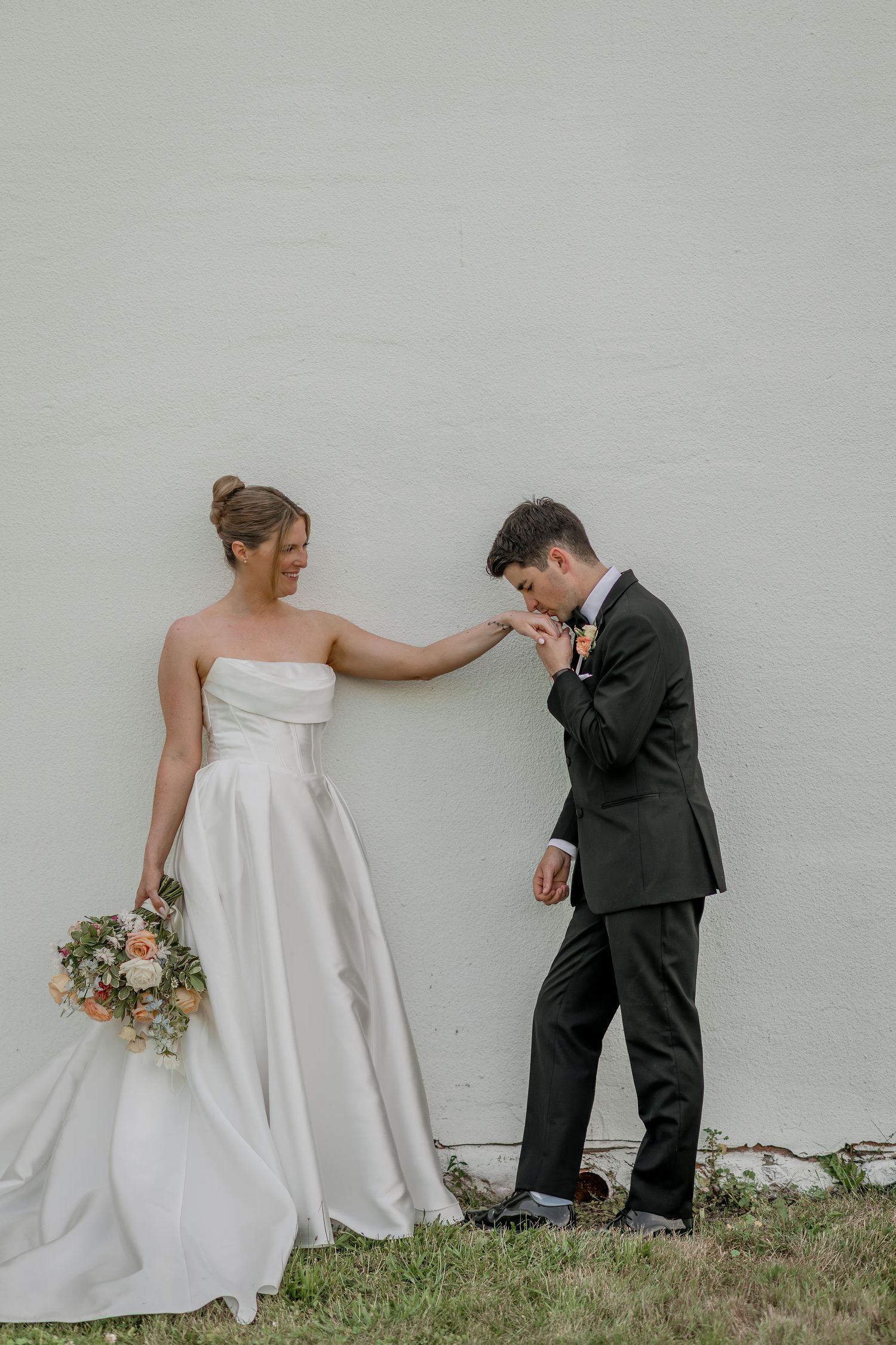 Artistic wedding couple poses playfully against white wall outdoors.