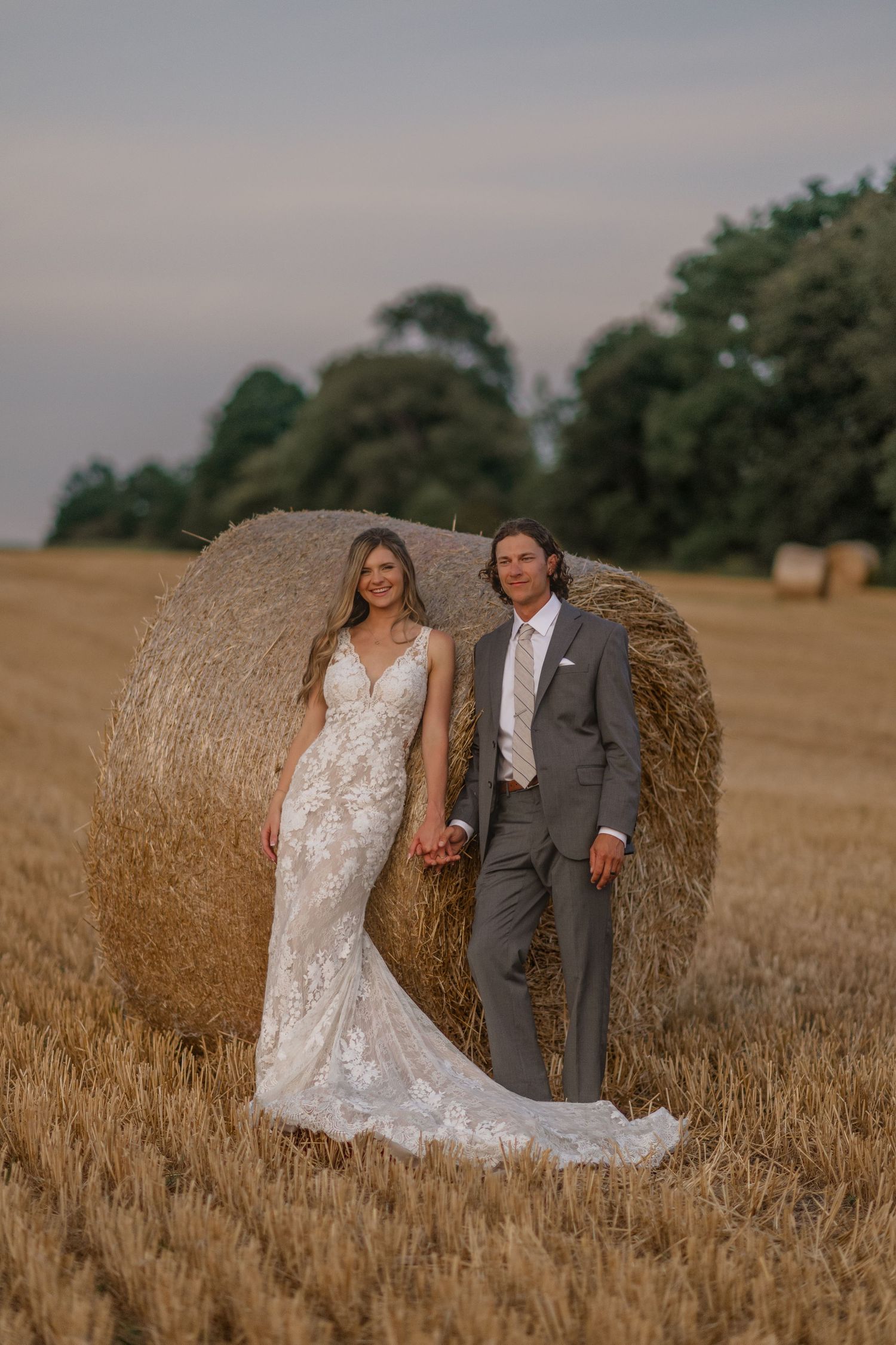 A wedding couple poses together against a large hay bale in a golden wheat field at sunset.