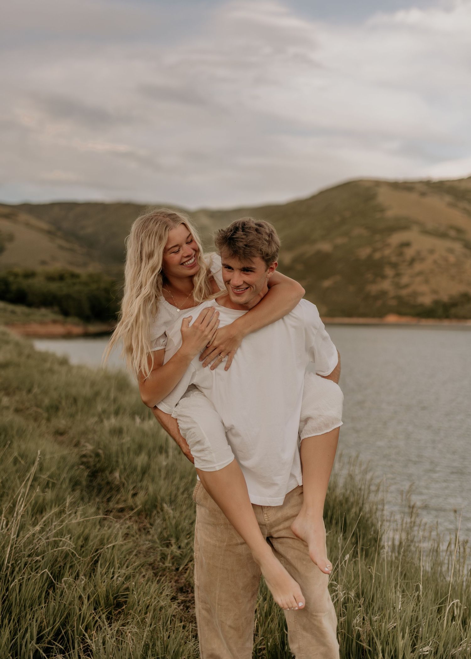 A couple in white shirts embraces lovingly by a scenic lake surrounded by rolling hills on a cloudy day.