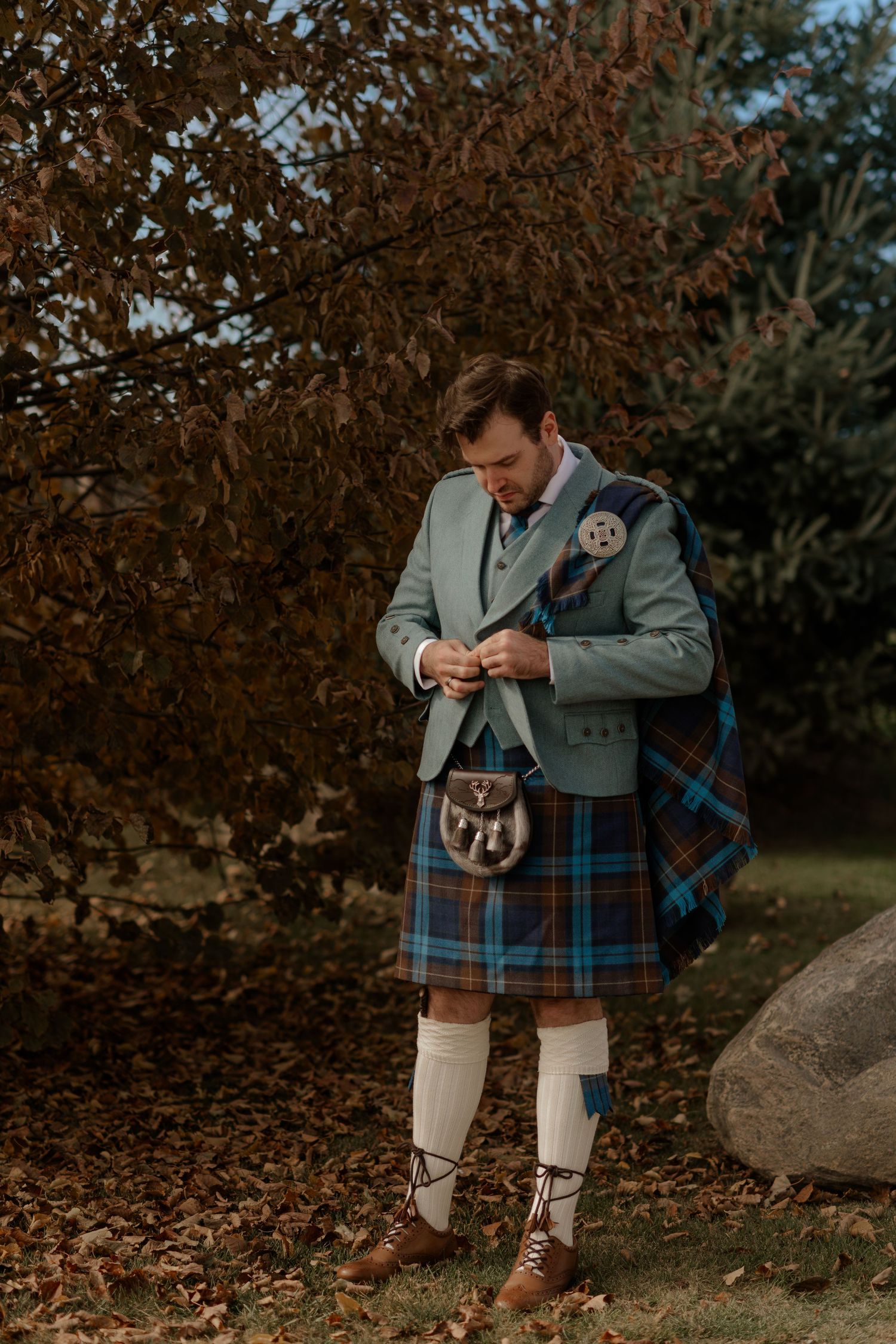 Traditional Scottish bagpiper in tartan kilt and gray jacket performs outdoors among autumn foliage.