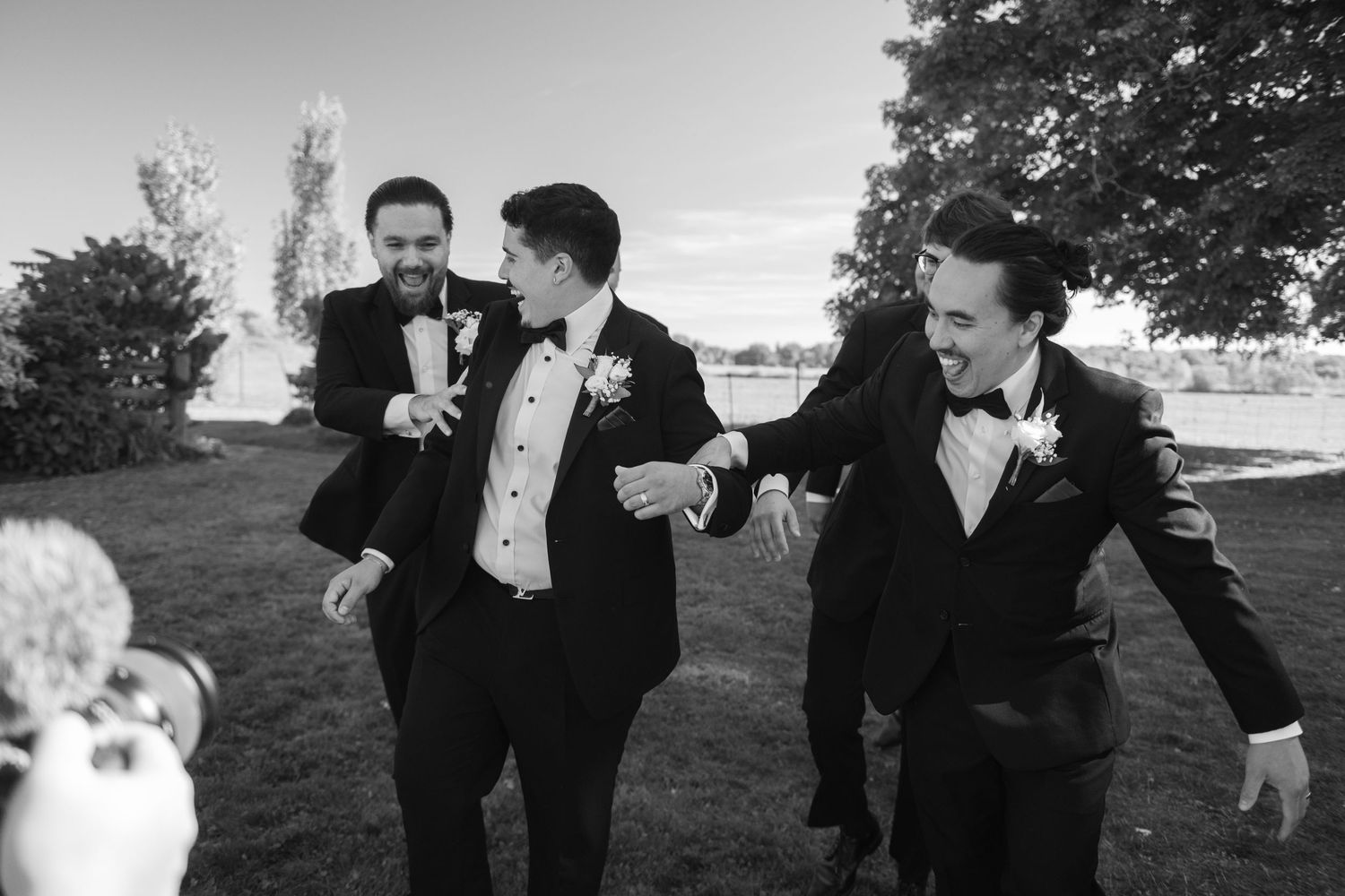 Three people in formal tuxedos laugh and share a moment together outdoors in black and white photography.