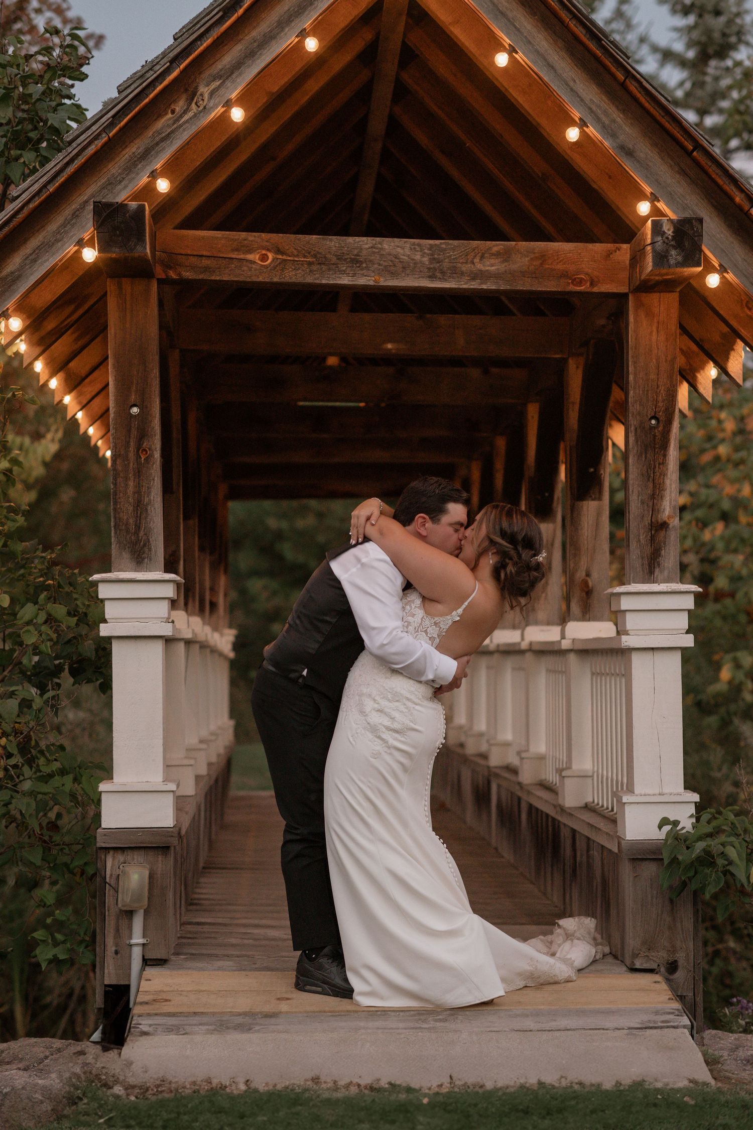Romantic couple shares intimate dance moments under a rustic wooden pavilion decorated with string lights at sunset.