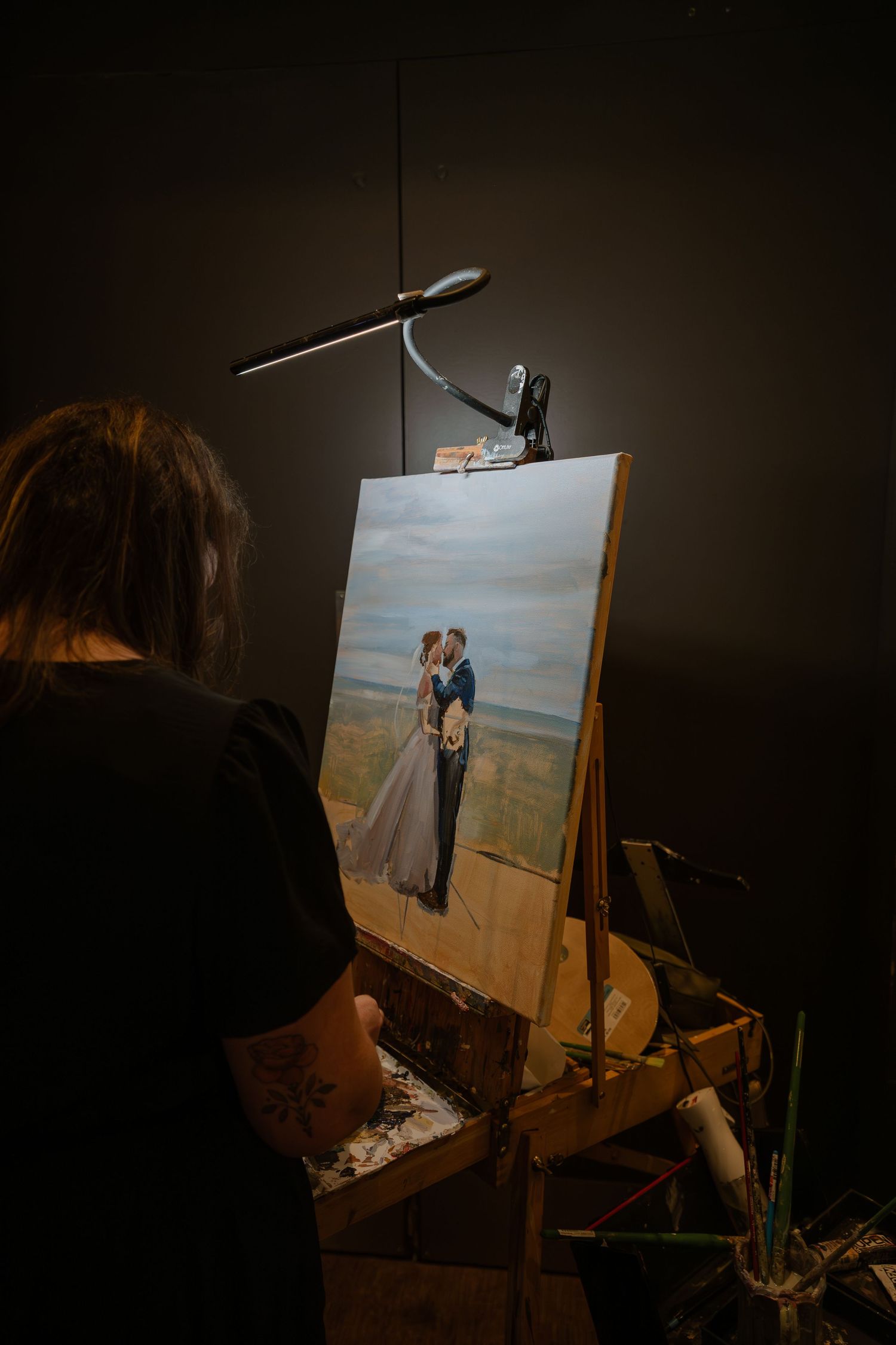 An artist works at an easel in a dark studio, painting what appears to be a landscape portrait.