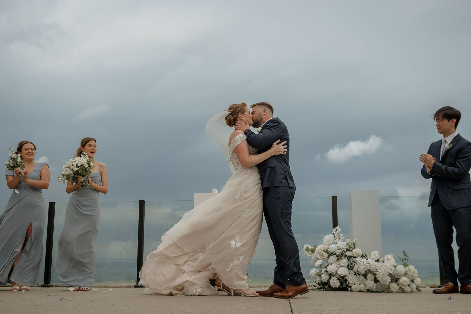 A wedding couple shares romantic moments on a pier with dramatic cloudy skies in the background.