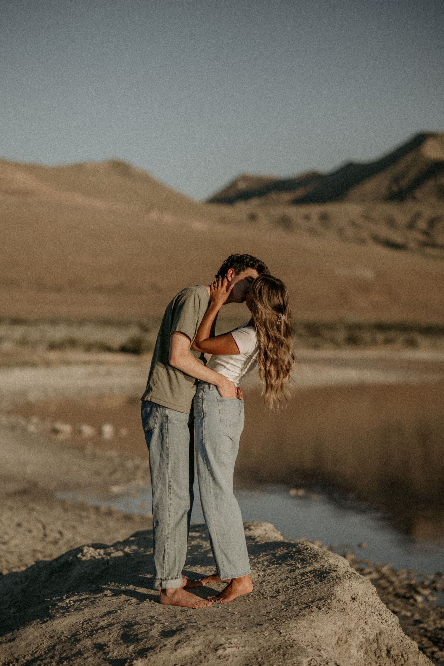 Two people share an intimate moment while standing on rocks near a desert lake with mountains in the background.