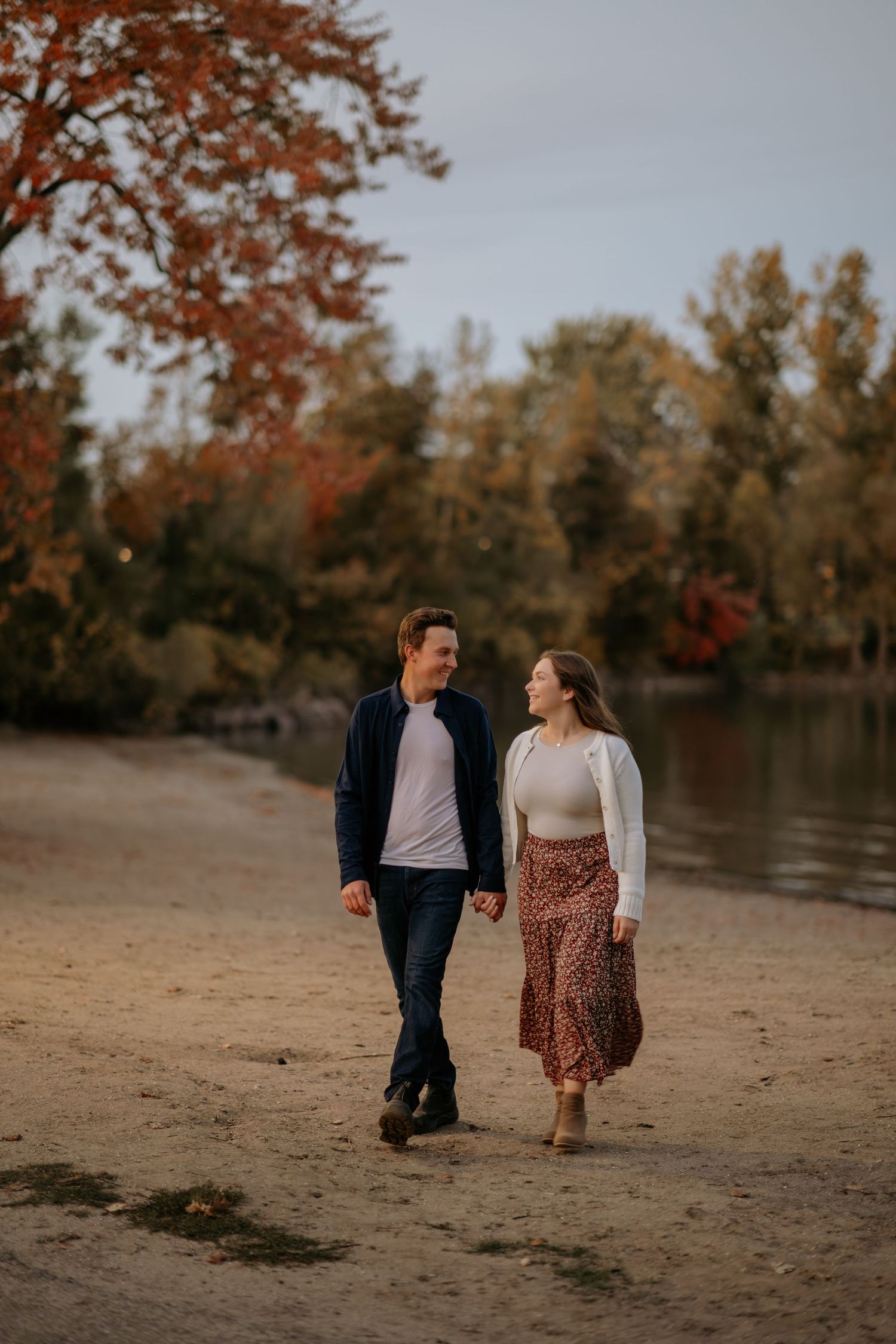Romantic couple walking on a beach at sunset with autumn trees in the background.